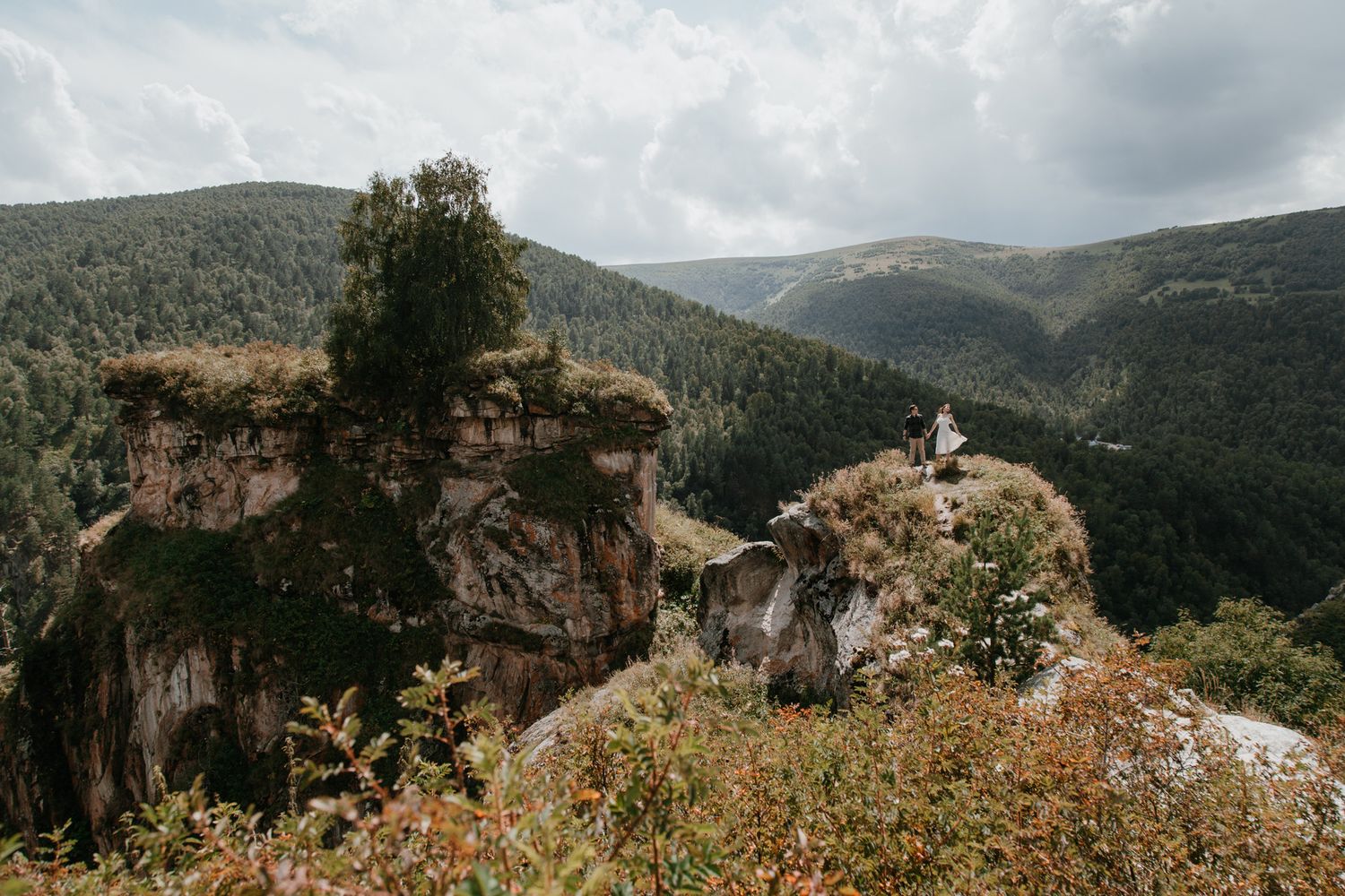A dramatic cliffside overlook with mountain ranges and forests stretching into the distance on a cloudy day.