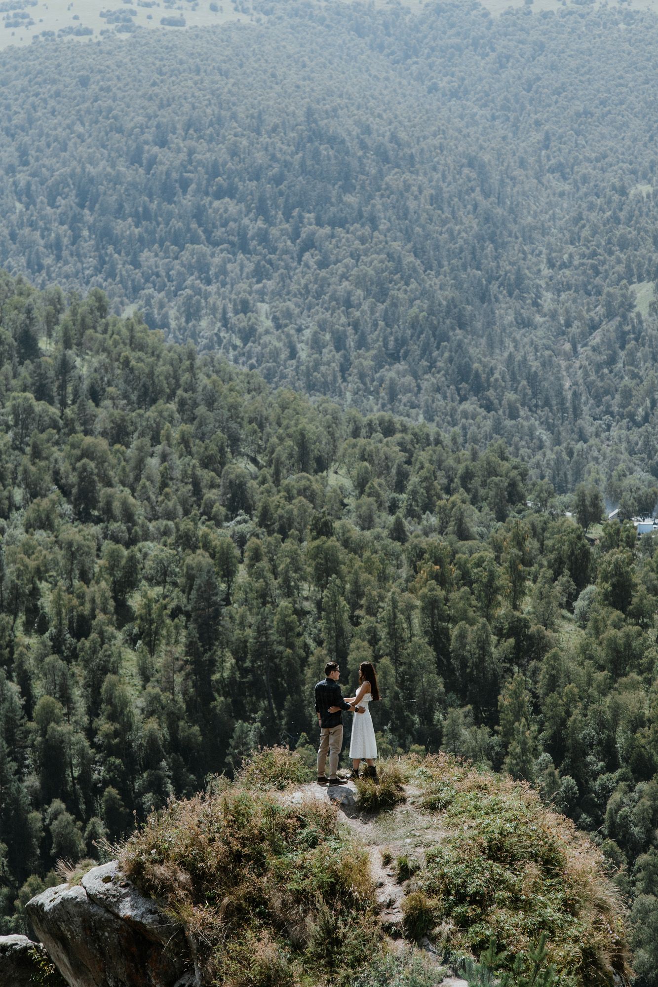 A couple stands on a dramatic cliffside overlook surrounded by dense forest and mountain views.
