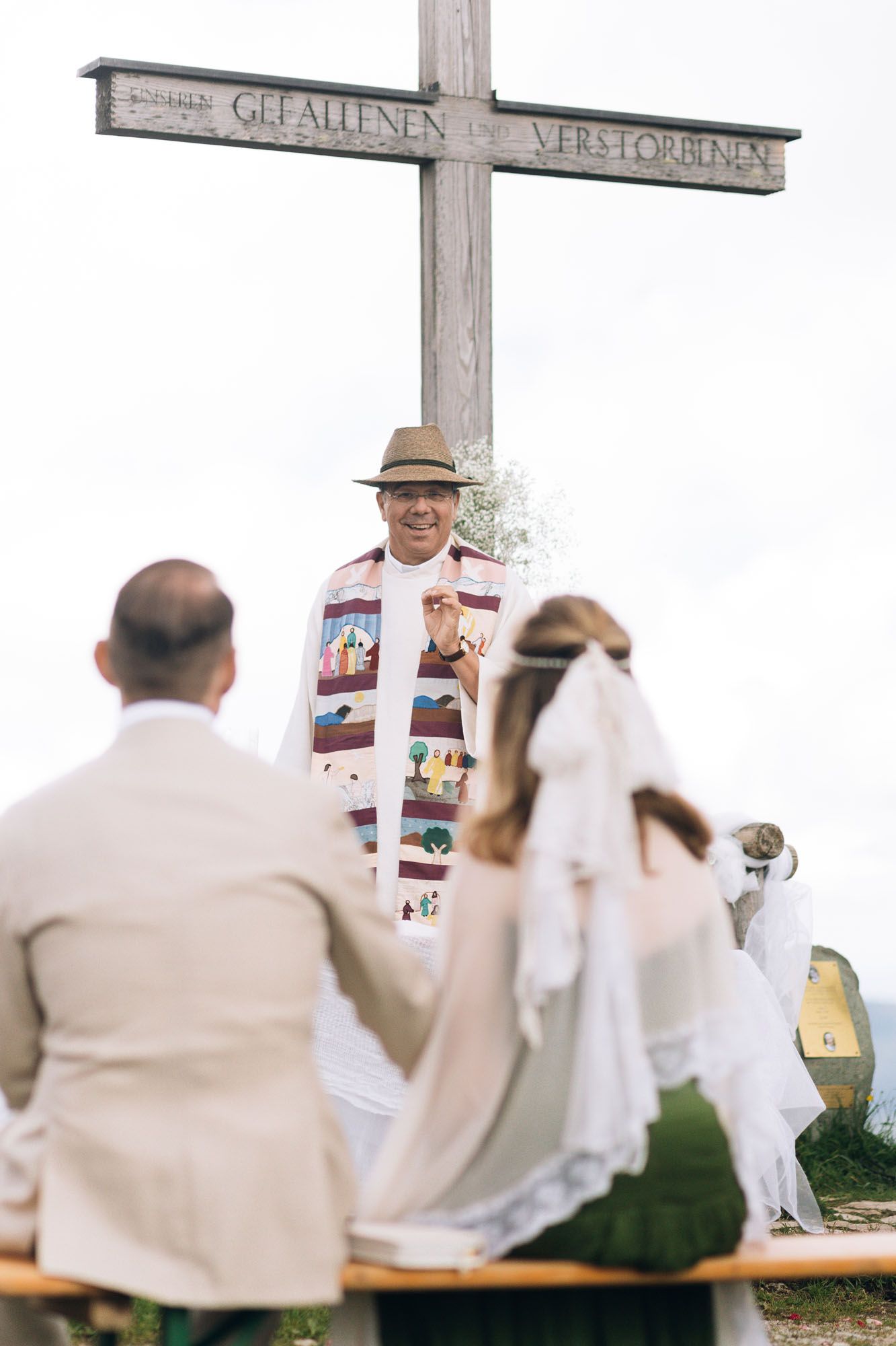 An outdoor wedding ceremony scene with a large wooden cross and people seated on benches.