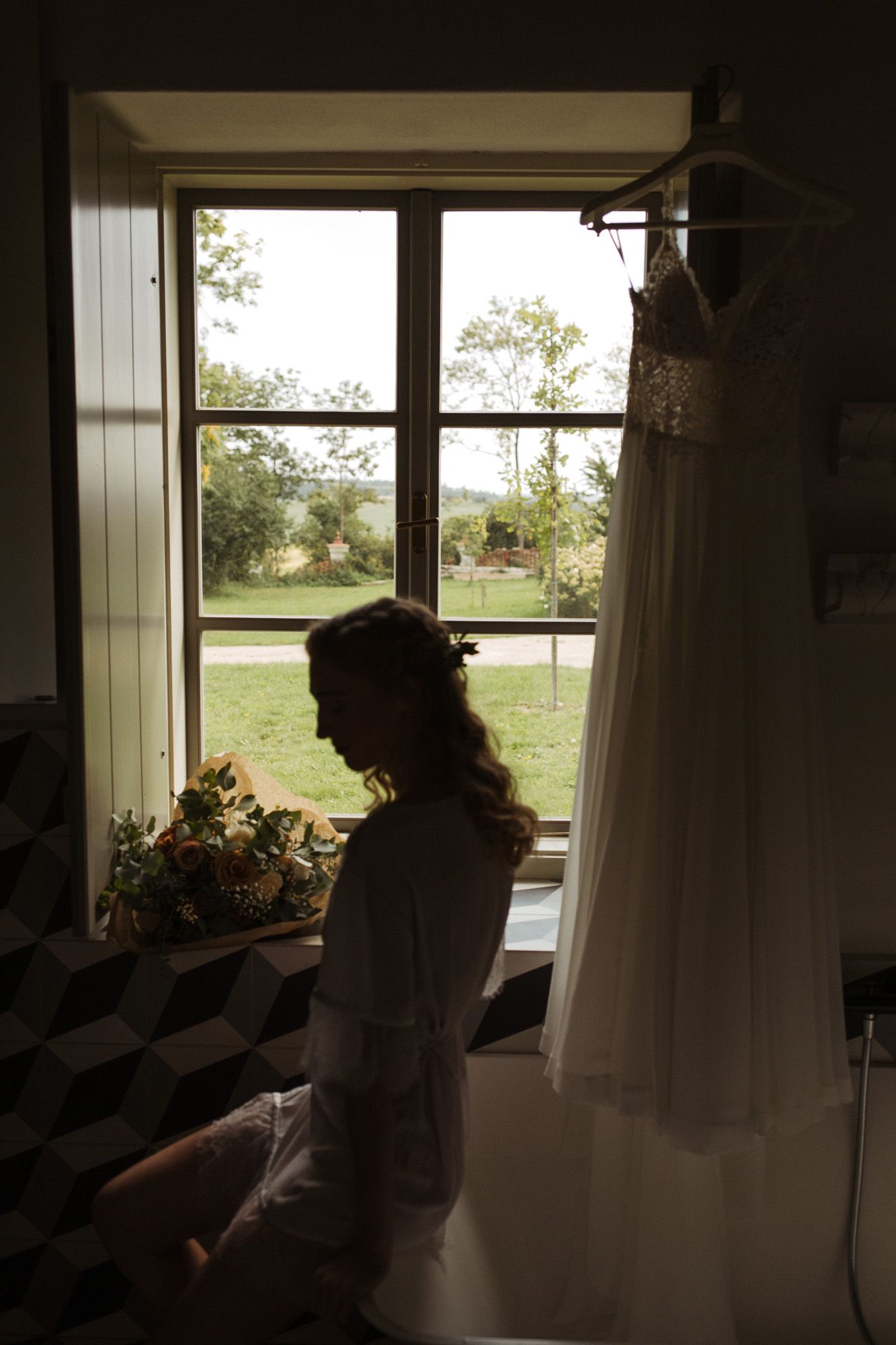 Silhouette by window with wedding dress hanging nearby in natural light.