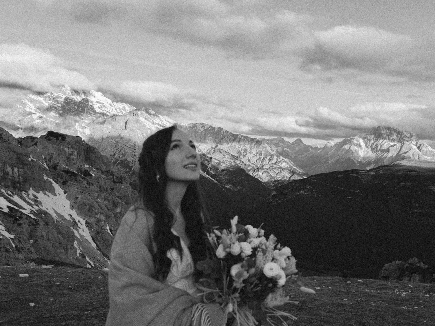 Black and white portrait of a mountain bride holding flowers with dramatic peaks in background.