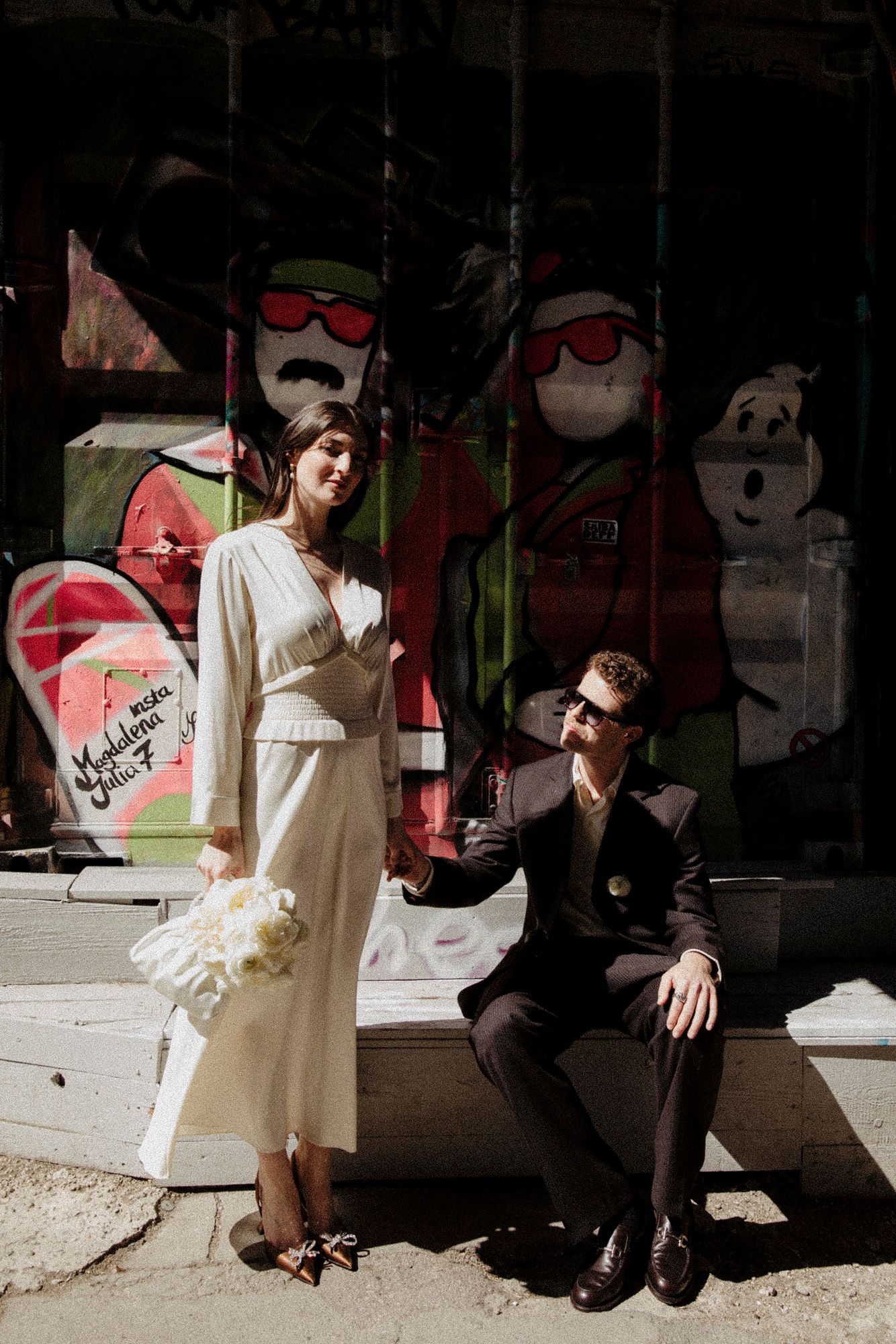 Couple in vintage-inspired wedding attire poses against graffiti wall with dramatic shadows and lighting.