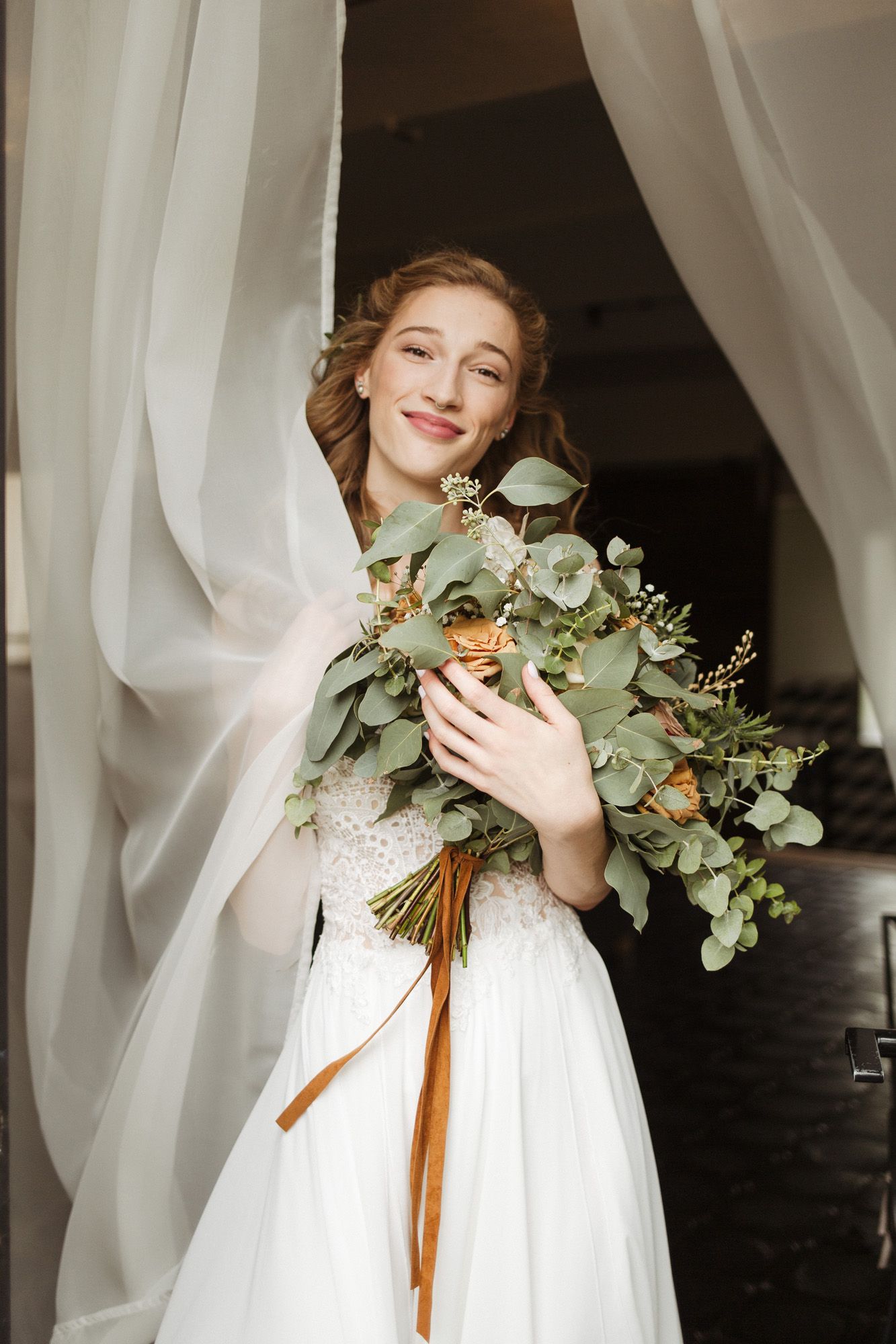 Bridal portrait with eucalyptus and peach rose bouquet against white curtains.