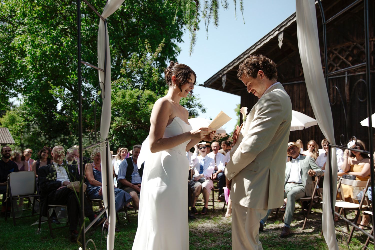 Wedding ceremony taking place under a decorated arch with gathered guests in a garden setting.