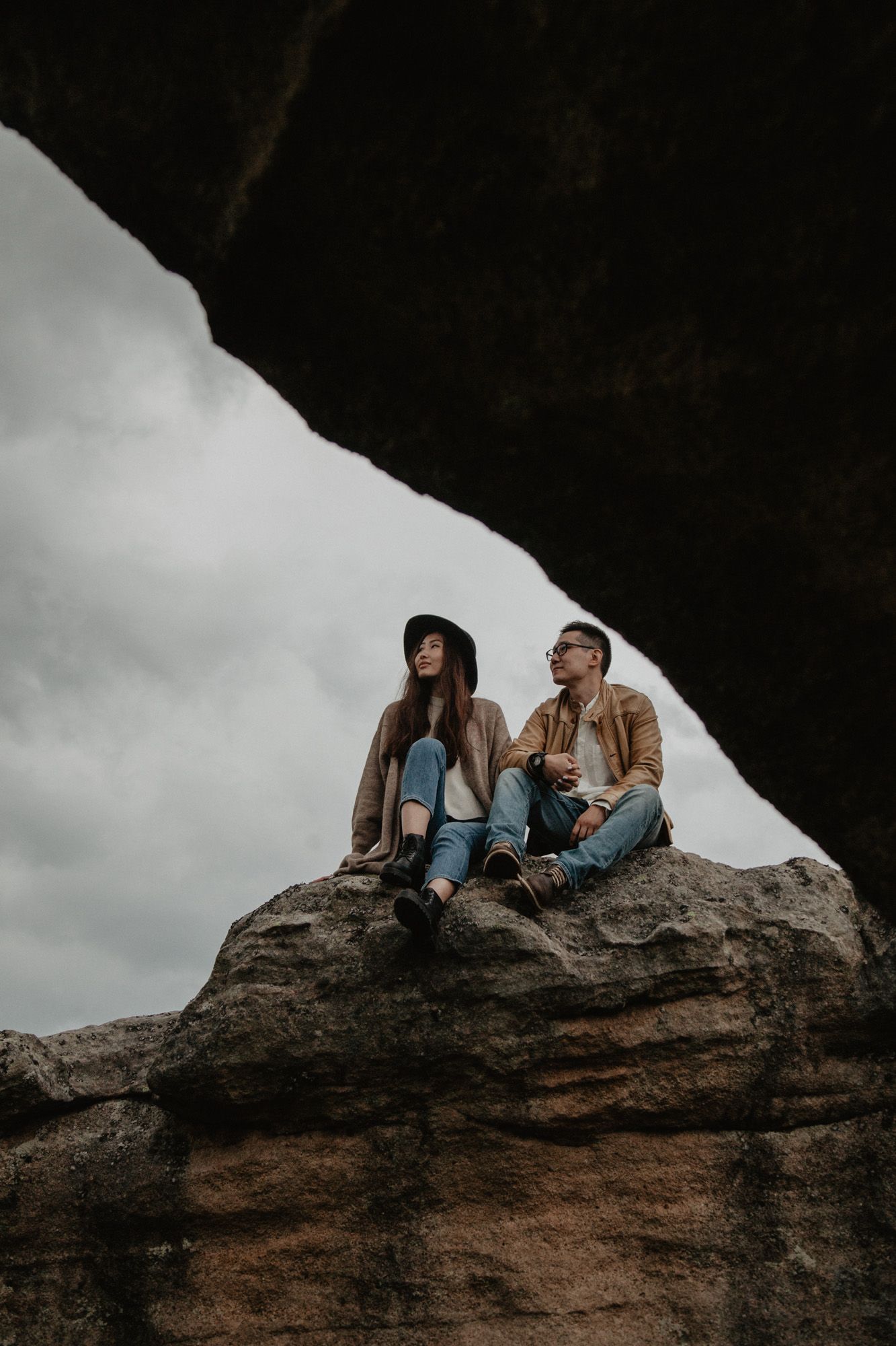 Two people sitting together on a dramatic rock formation against cloudy skies.