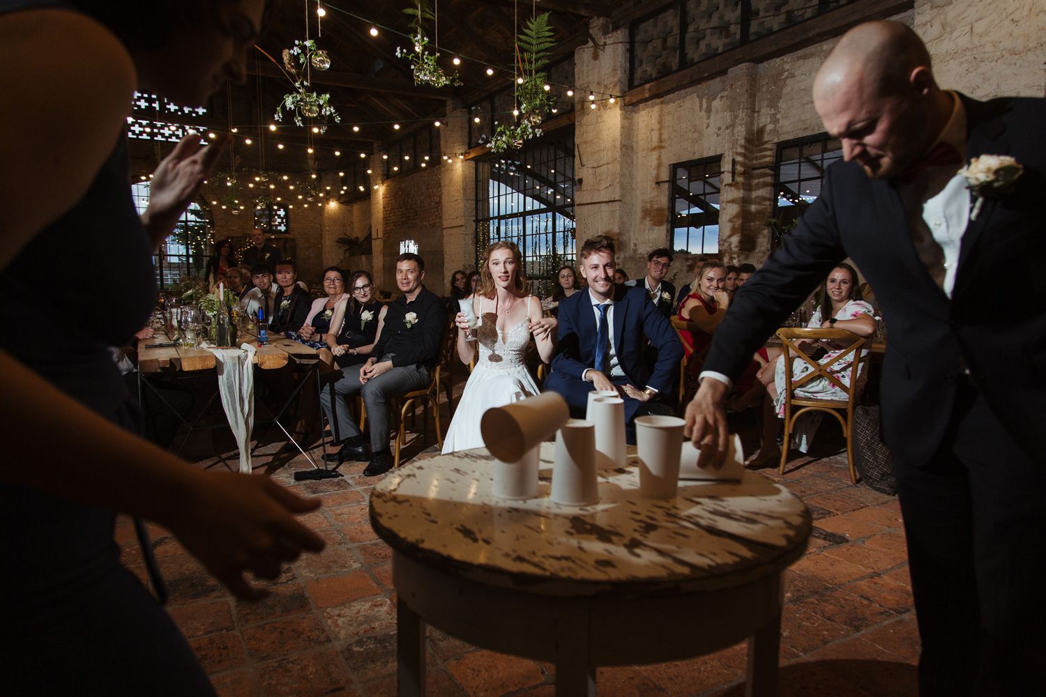 Indoor wedding reception with guests gathered around wooden tables in a rustic brick venue.