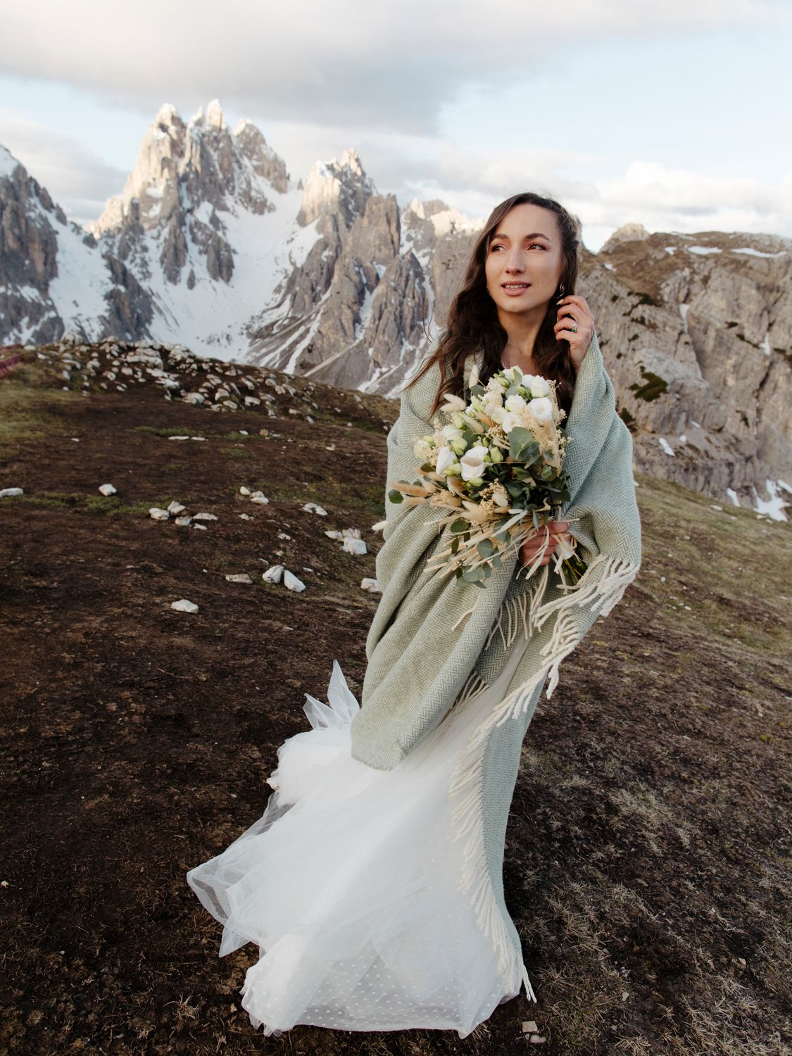 A bride in a flowing white dress holds a bouquet against majestic mountain peaks at sunset.