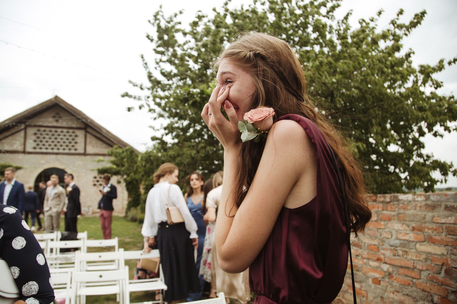 Outdoor wedding ceremony setup with guests seated on white chairs near a tree-lined brick venue.