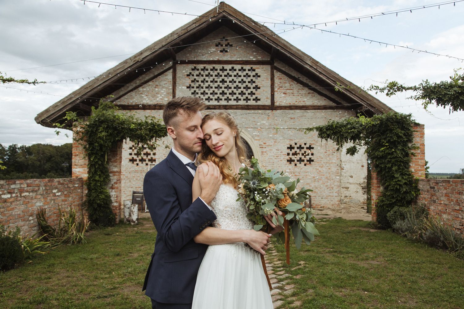 A romantic wedding portrait in front of a vintage barn with decorative lattice details and string lights.