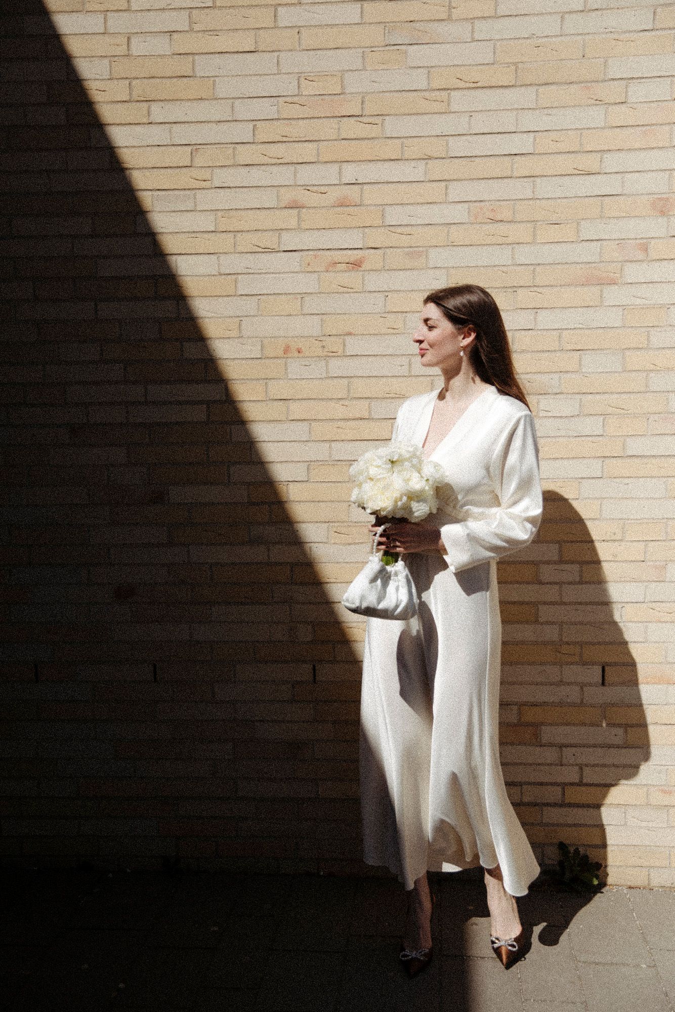 A bride in a sleek white dress poses against a brick wall with dramatic lighting and shadows.