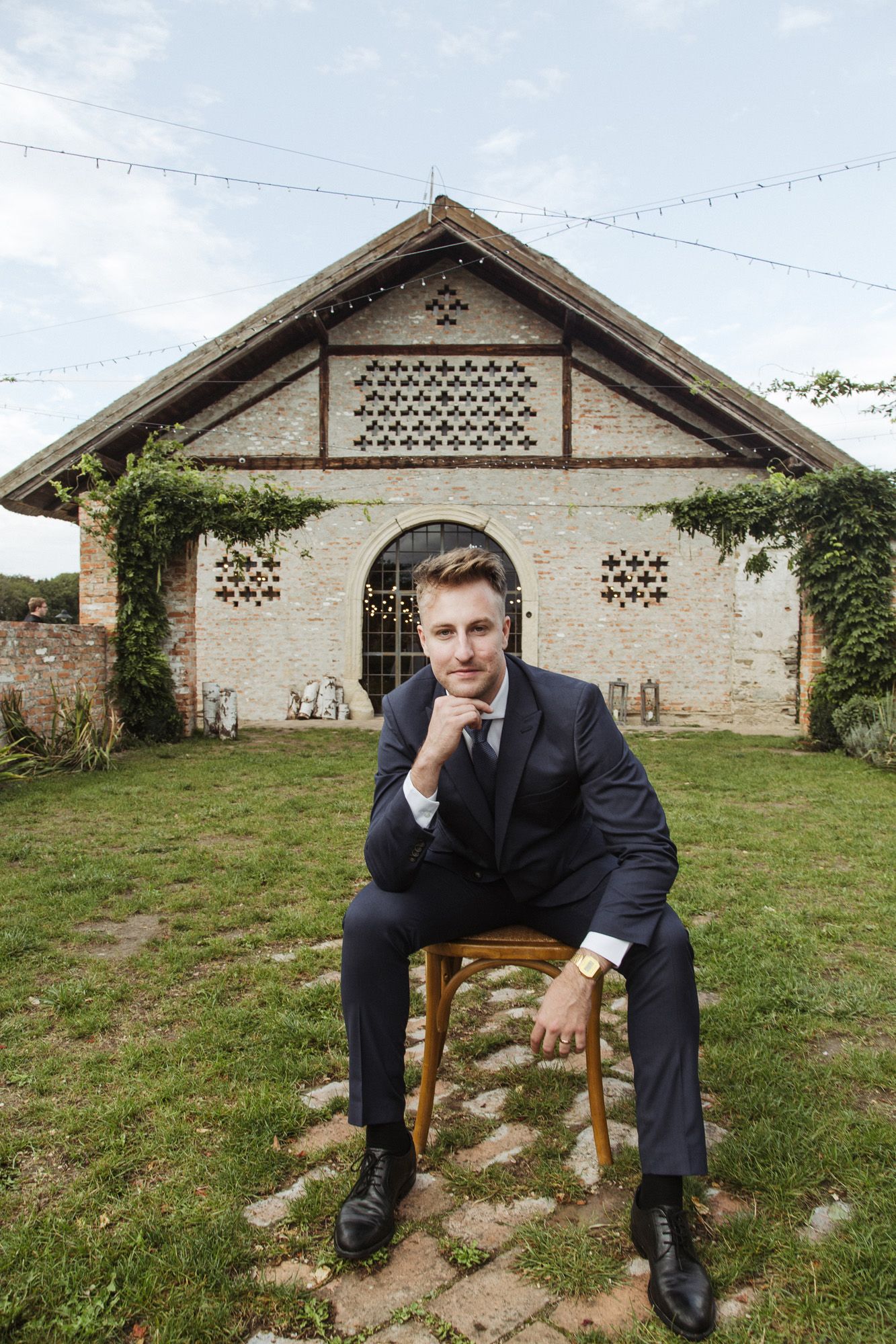 Groom in navy suit sits on wooden chair against rustic building backdrop.