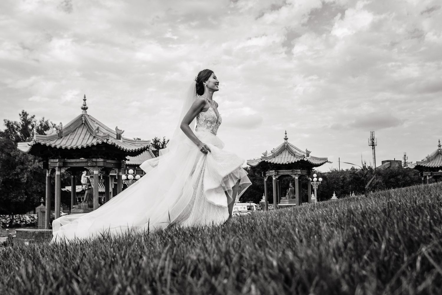 Bride in flowing white dress poses in garden with Asian pagodas in background.