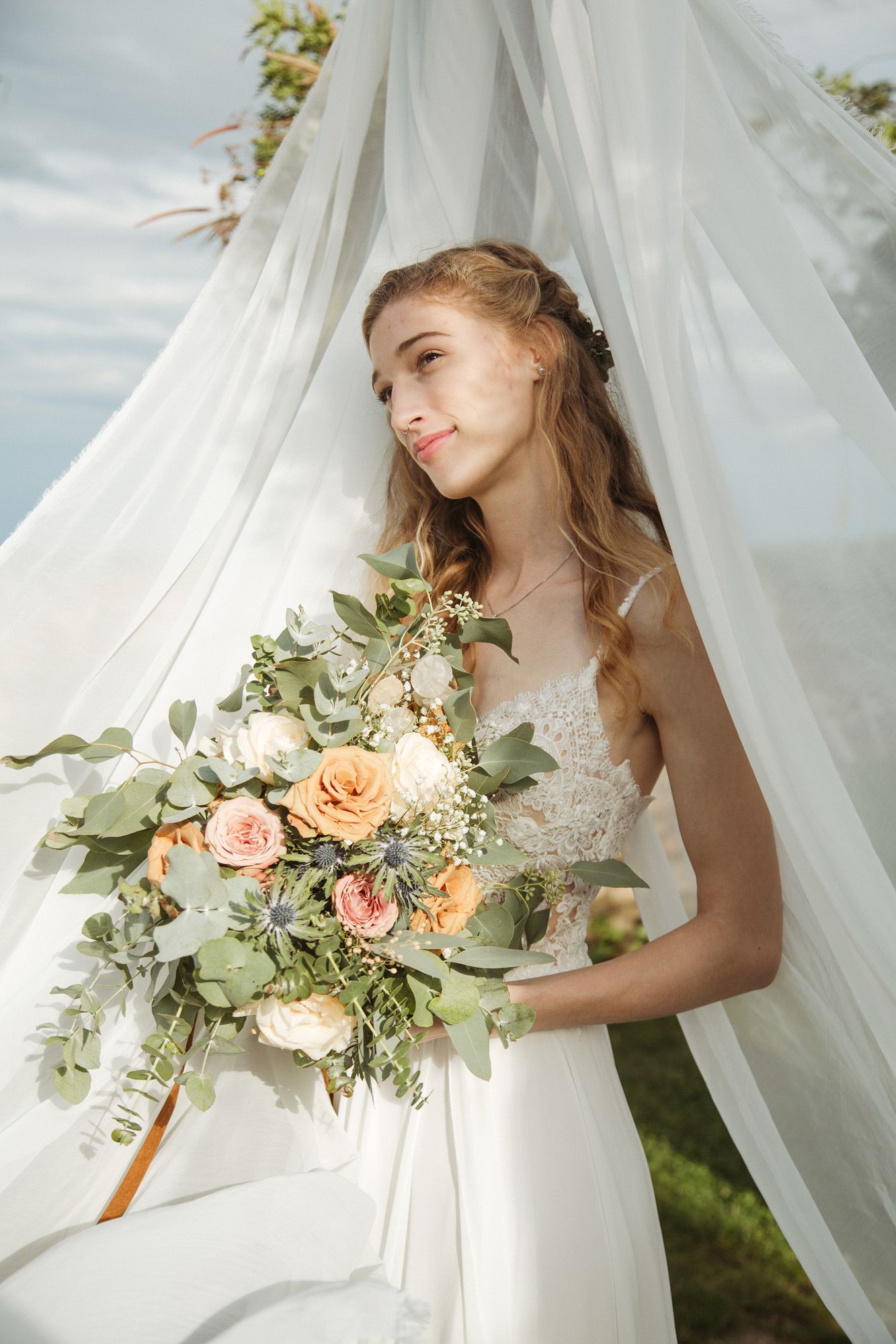 A bride in white lace holds a peach and green bouquet against flowing sheer fabric outdoors.