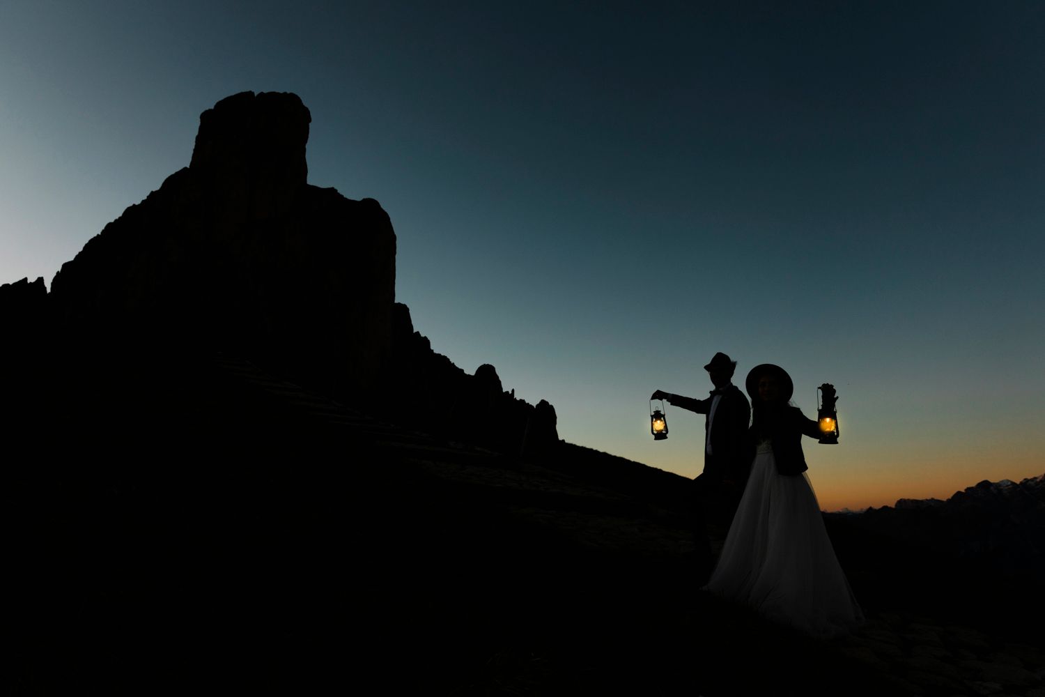 Silhouettes hold lanterns against a dramatic mountain sunset backdrop.