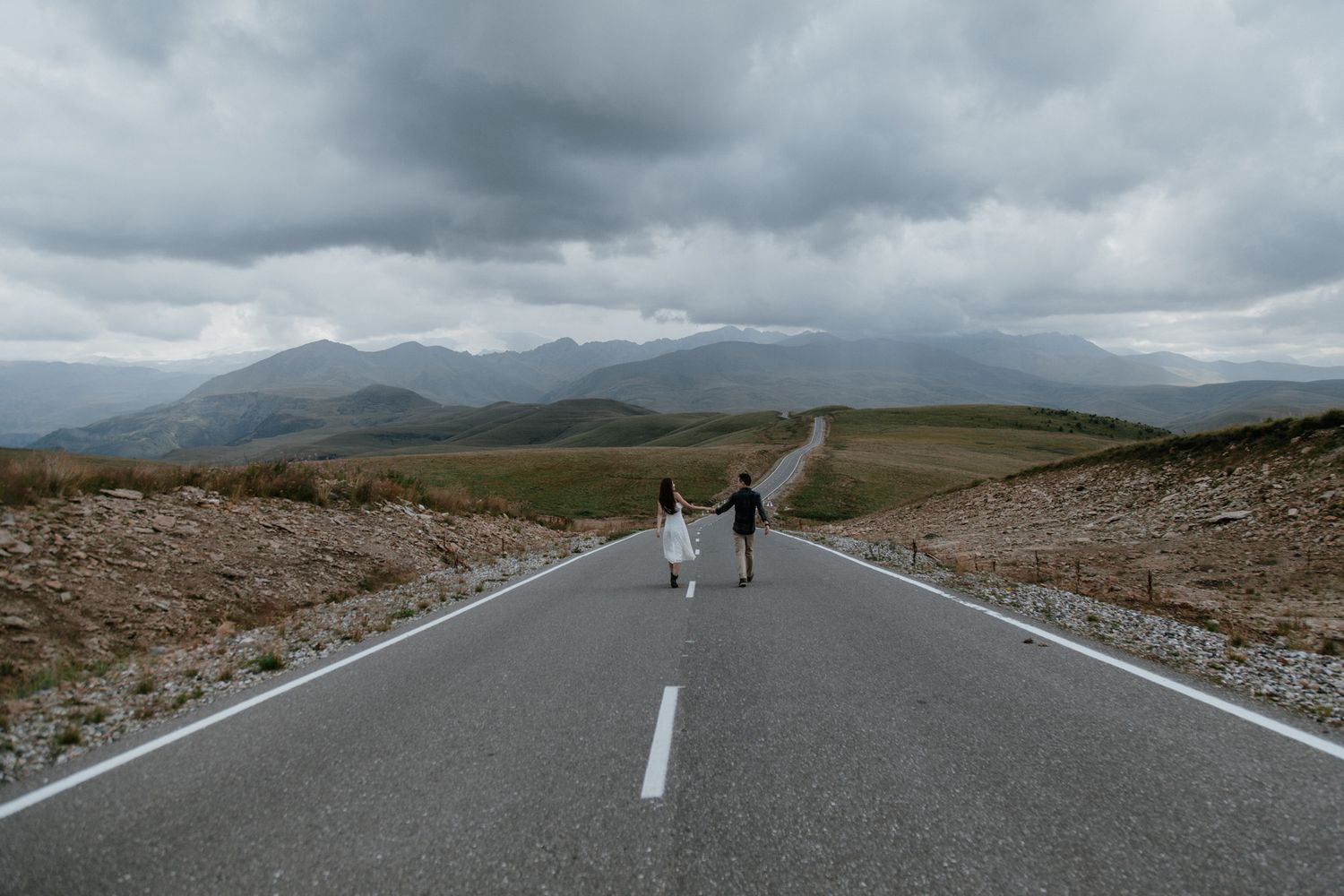 Two figures walking down a remote mountain road with dramatic cloudy skies and mountains in the distance.