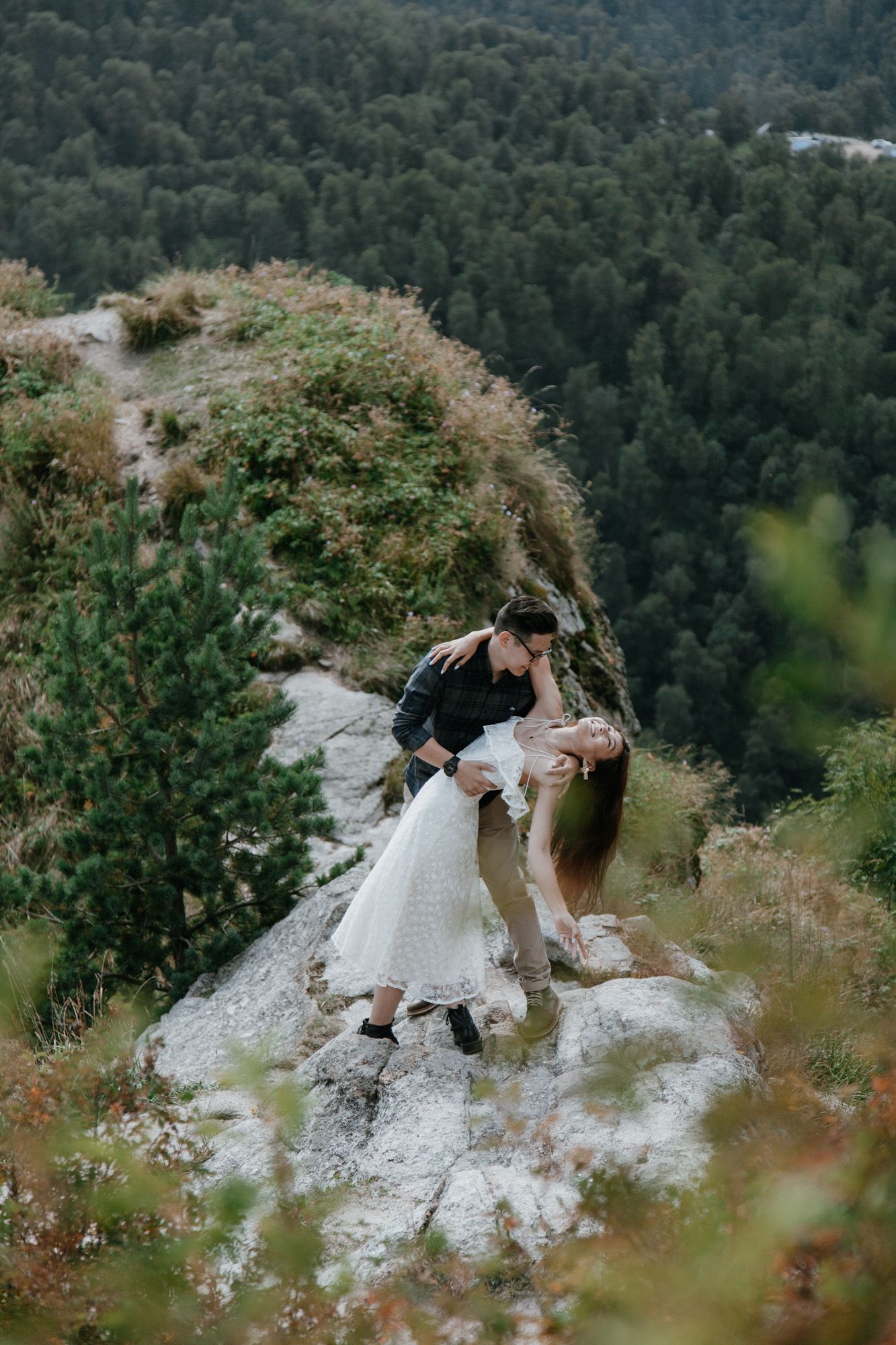 A romantic couple poses dramatically on a rocky cliff edge overlooking a forested valley.