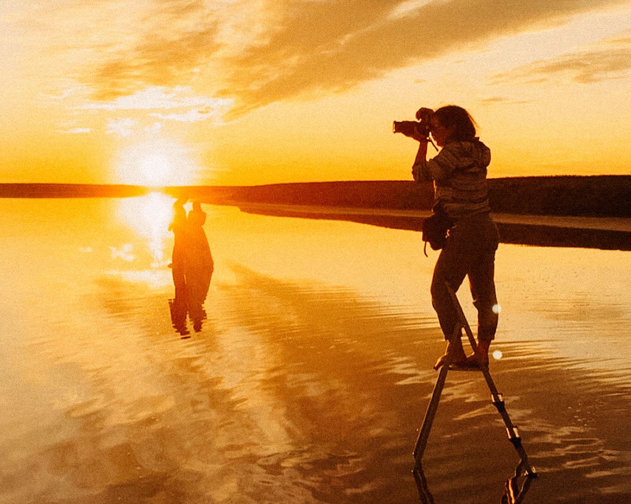 Photographer on stilts captures silhouettes against golden sunset on beach.