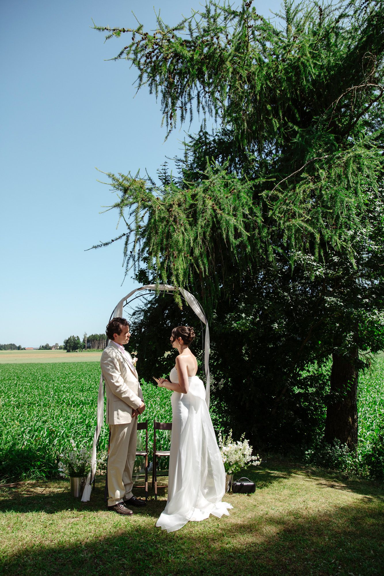 Sunlit outdoor wedding ceremony next to a green farm field under weeping willow trees.