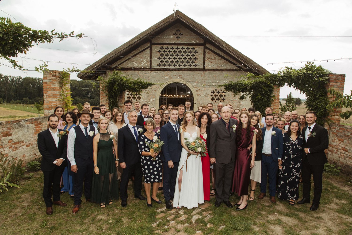 Wedding guests gather for group photo outside rustic brick venue with decorative lattice details.