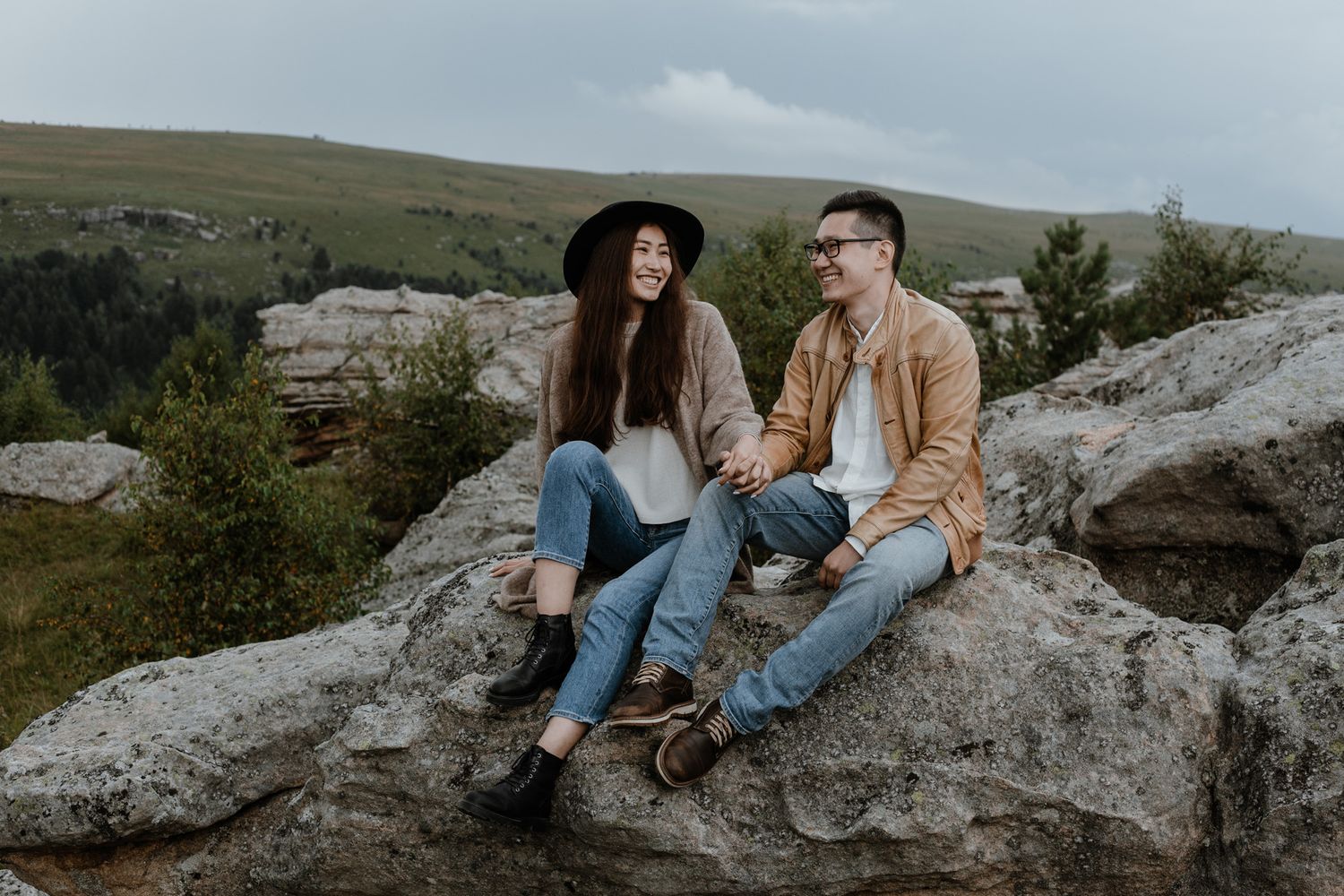 Couple sitting together on rocky outcrop overlooking scenic mountain landscape at sunset.