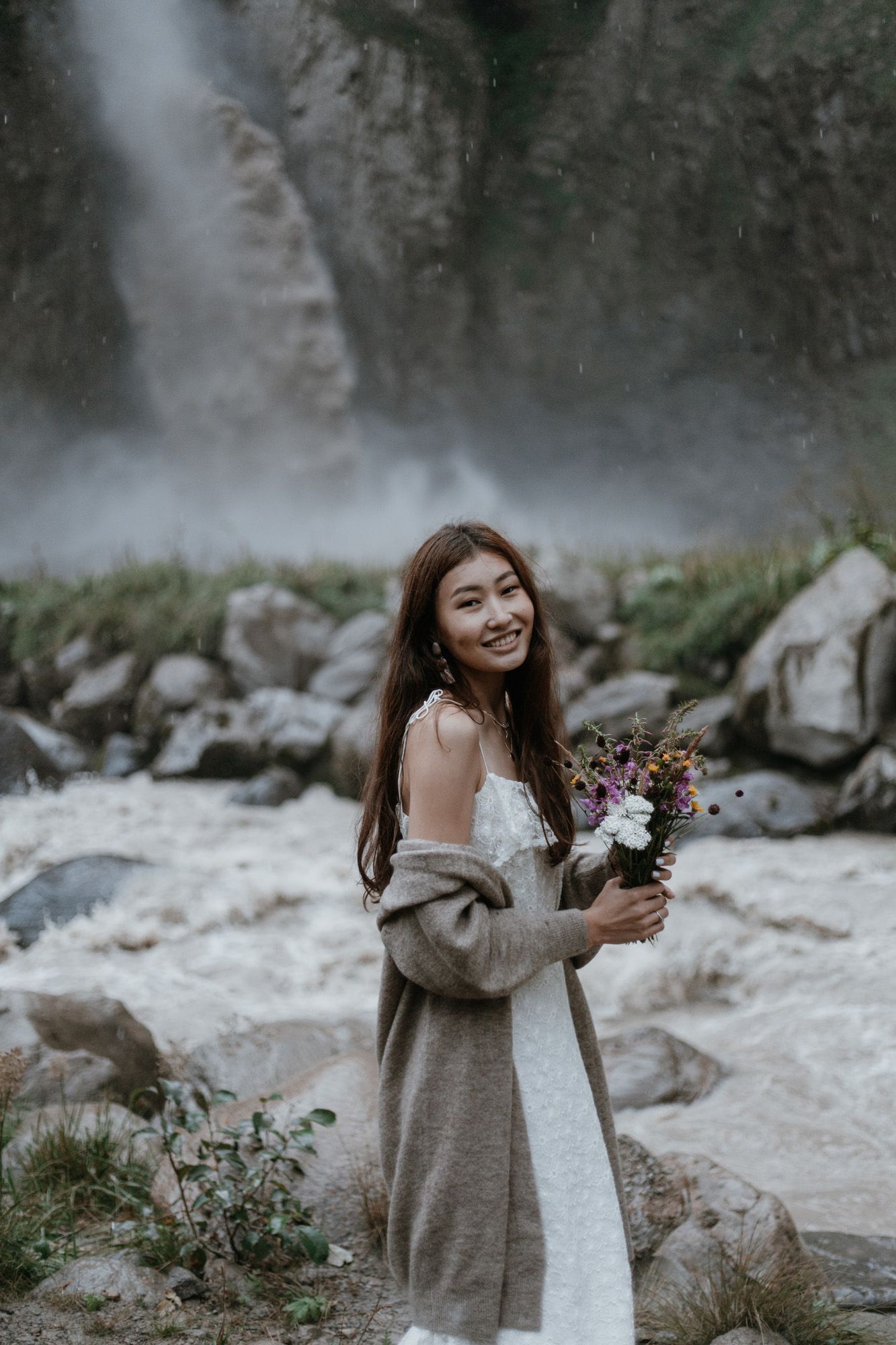 A figure in white holds flowers by a misty waterfall surrounded by rocky cliffs.