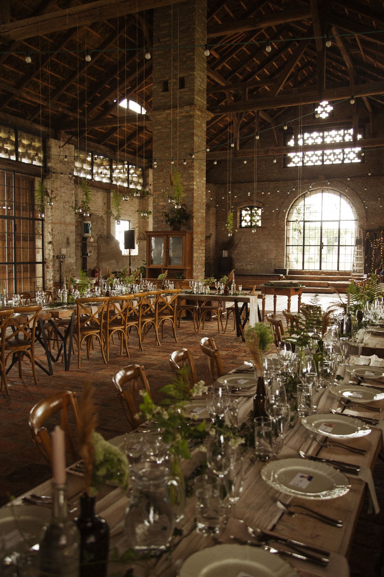 Rustic indoor wedding reception venue with long wooden tables and elegant place settings under exposed beams and natural lighting.