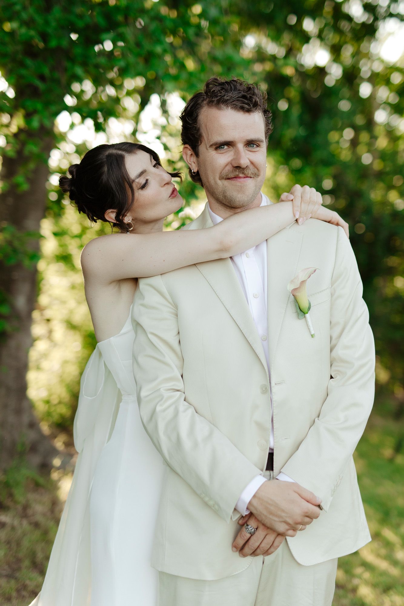 Romantic wedding couple in white attire embracing outdoors in a garden setting with soft natural lighting.
