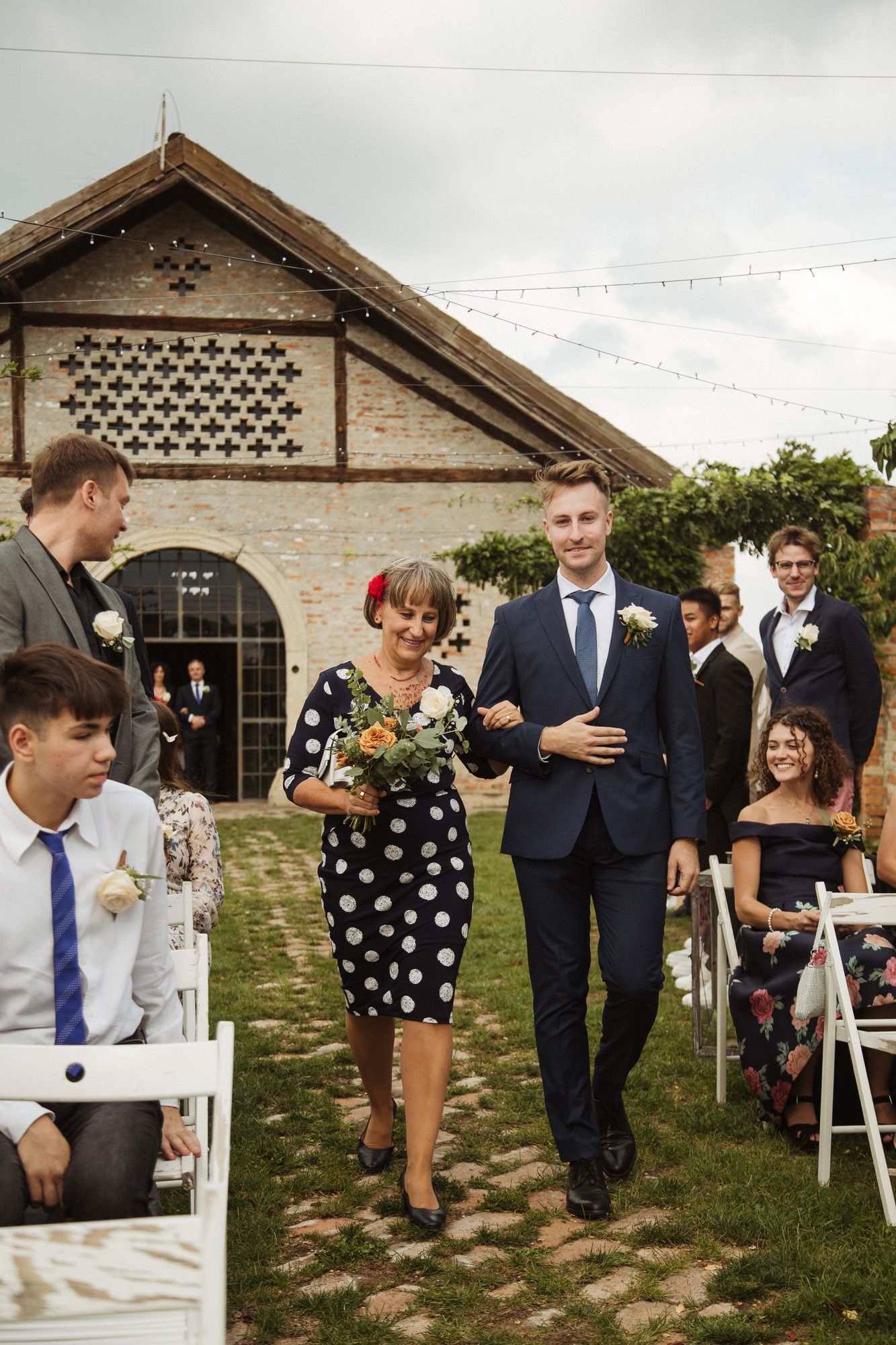 Wedding guests gather on a grassy lawn outside a charming brick venue with decorative lattice details.
