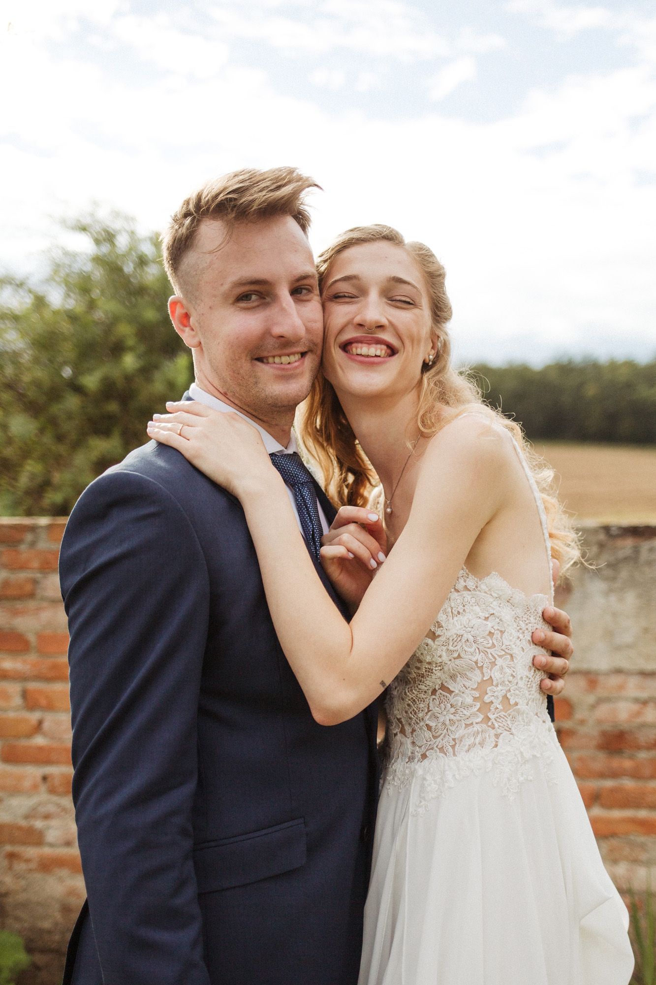 A joyful wedding couple embraces and laughs together against a sunny outdoor backdrop.