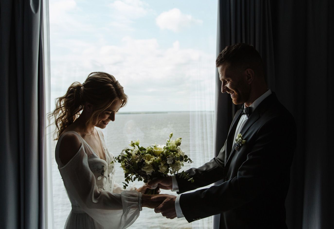 Silhouetted couple stands by window with wedding bouquet in hands during intimate wedding ceremony.