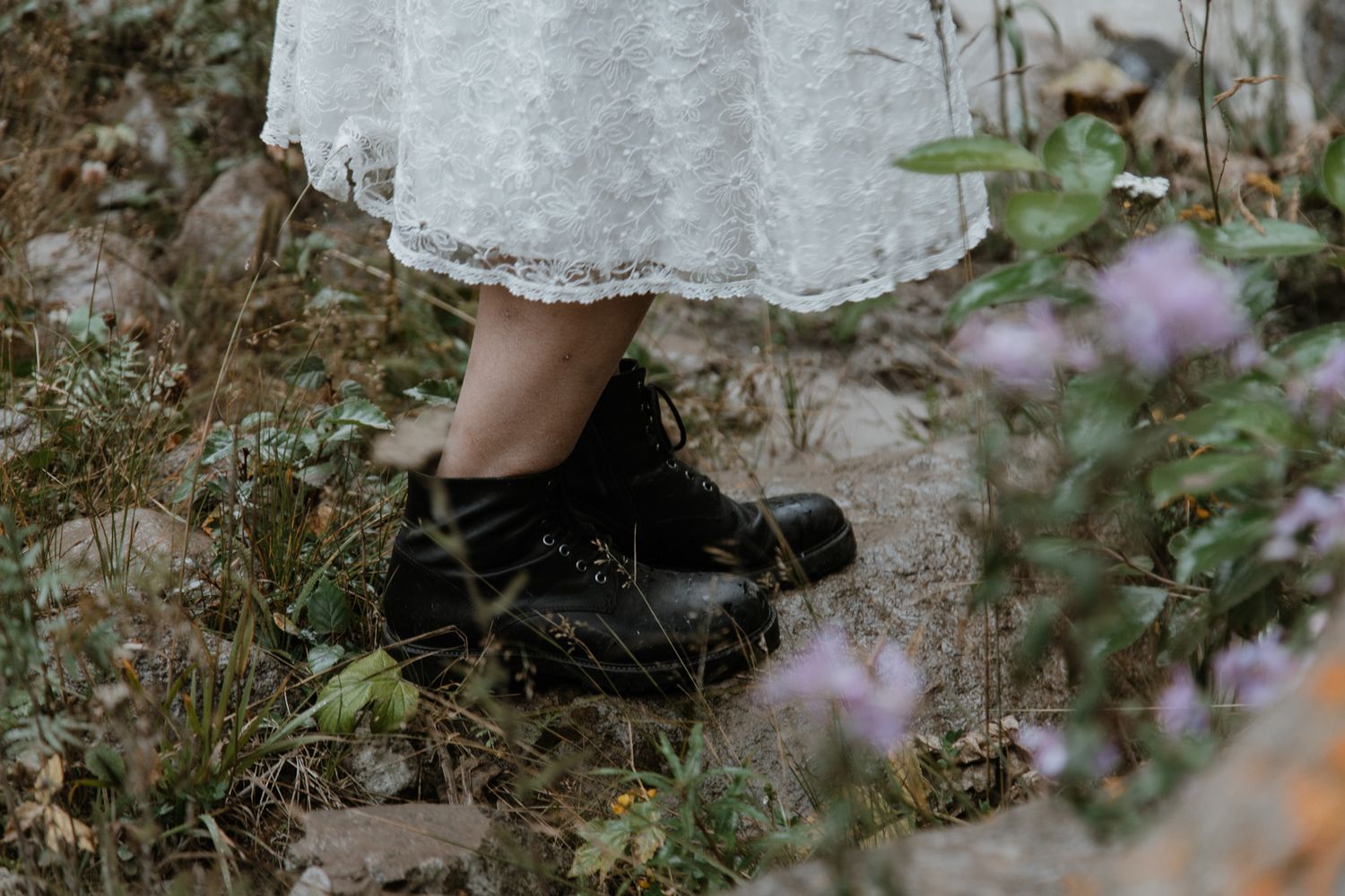 Close-up detail of black boots and lace dress hem against wildflowers.