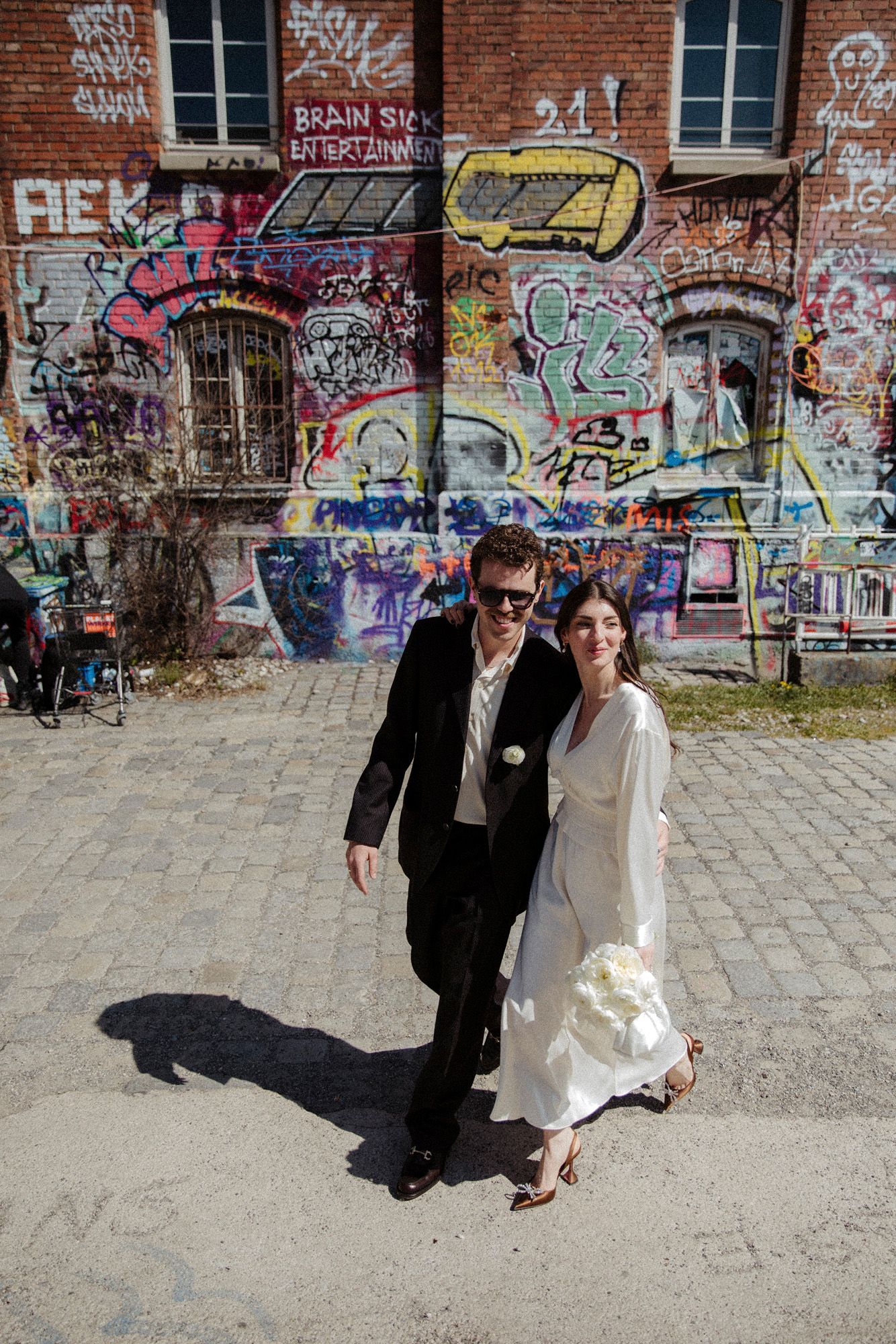 Wedding couple poses playfully in front of brick wall covered in colorful street art and graffiti.