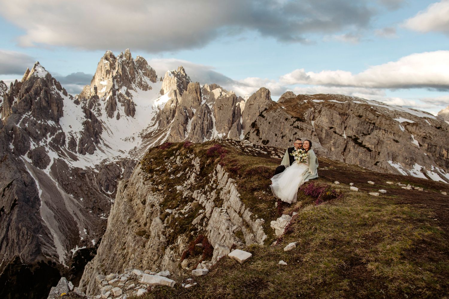 Dramatic mountain elopement photo with couple sitting on cliff edge overlooking rocky peaks.