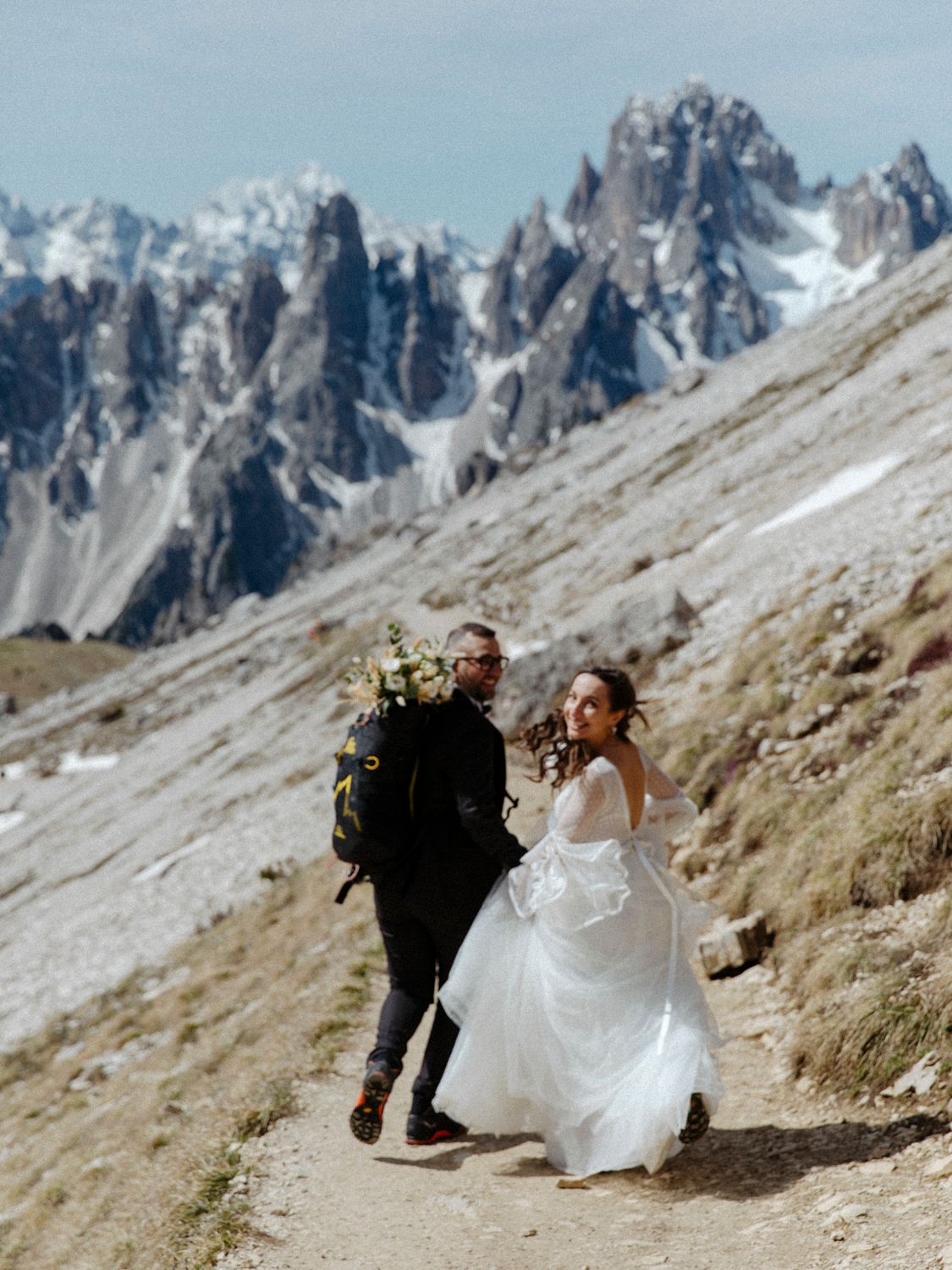 Wedding couple hiking along mountain trail with dramatic peaks in background.