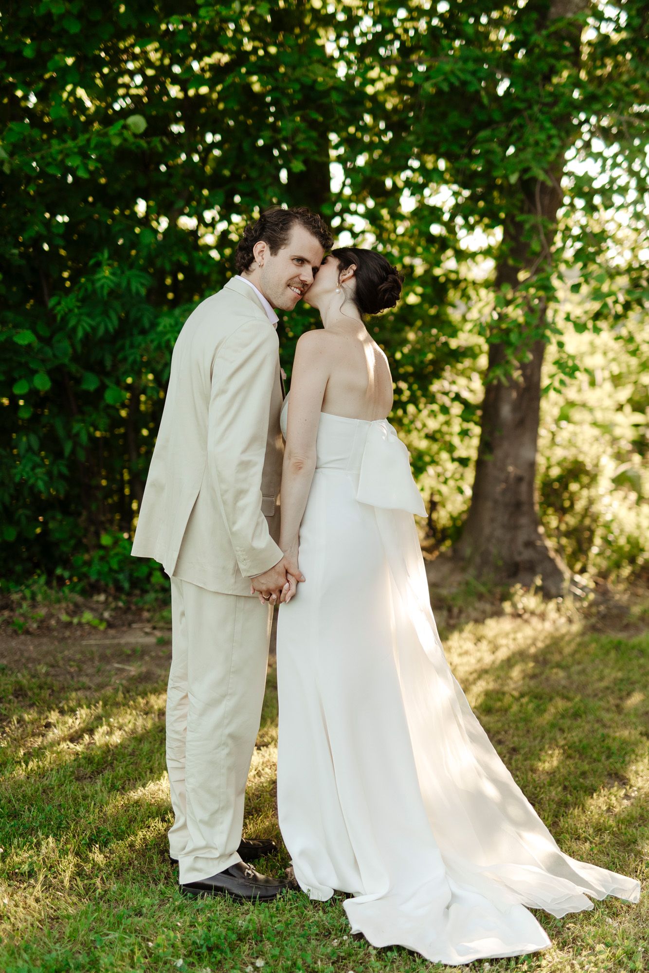 Newlyweds in elegant white attire share tender moments on a sunlit grassy hillside.