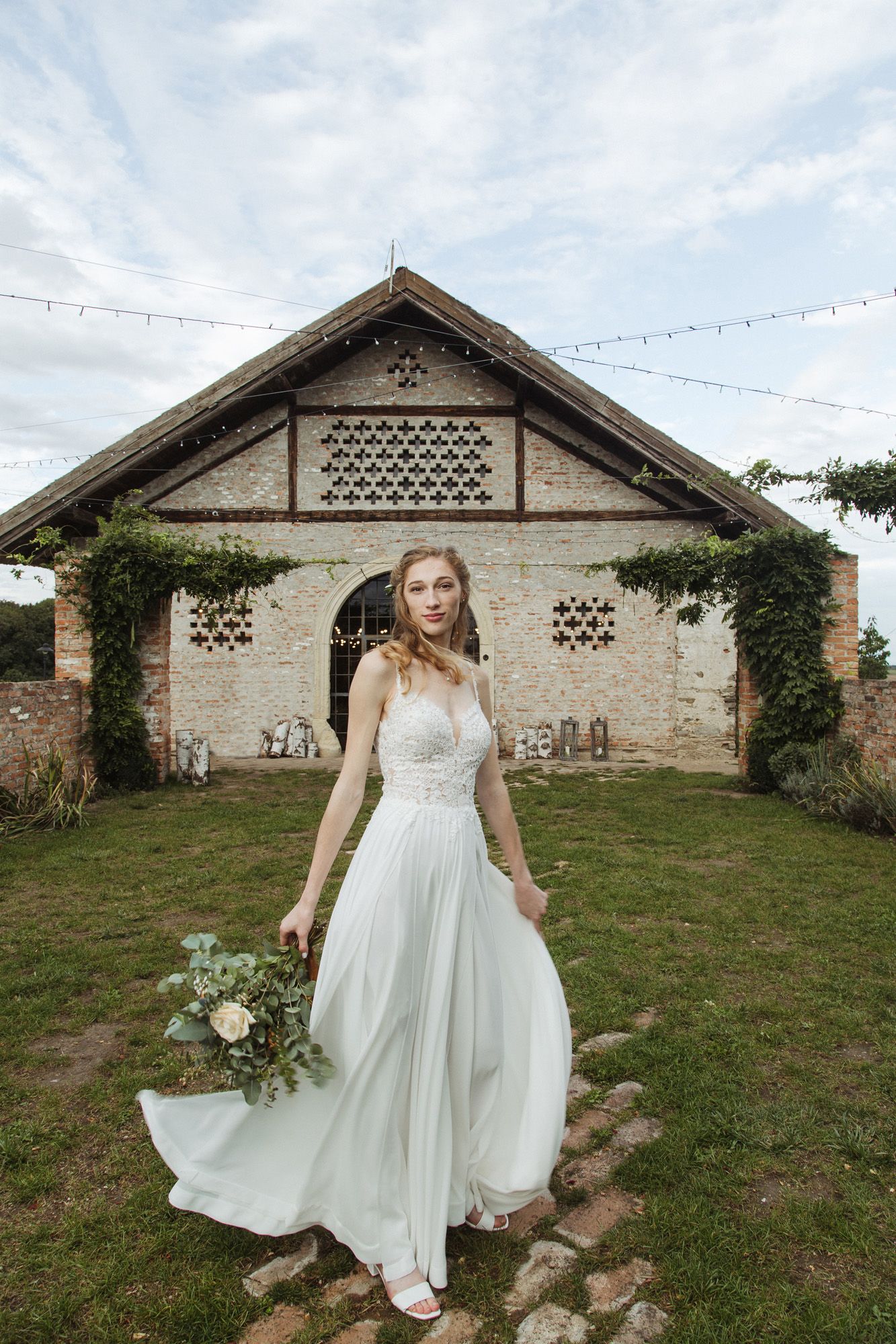 A flowing white wedding dress captured against a rustic brick barn with string lights and climbing ivy.
