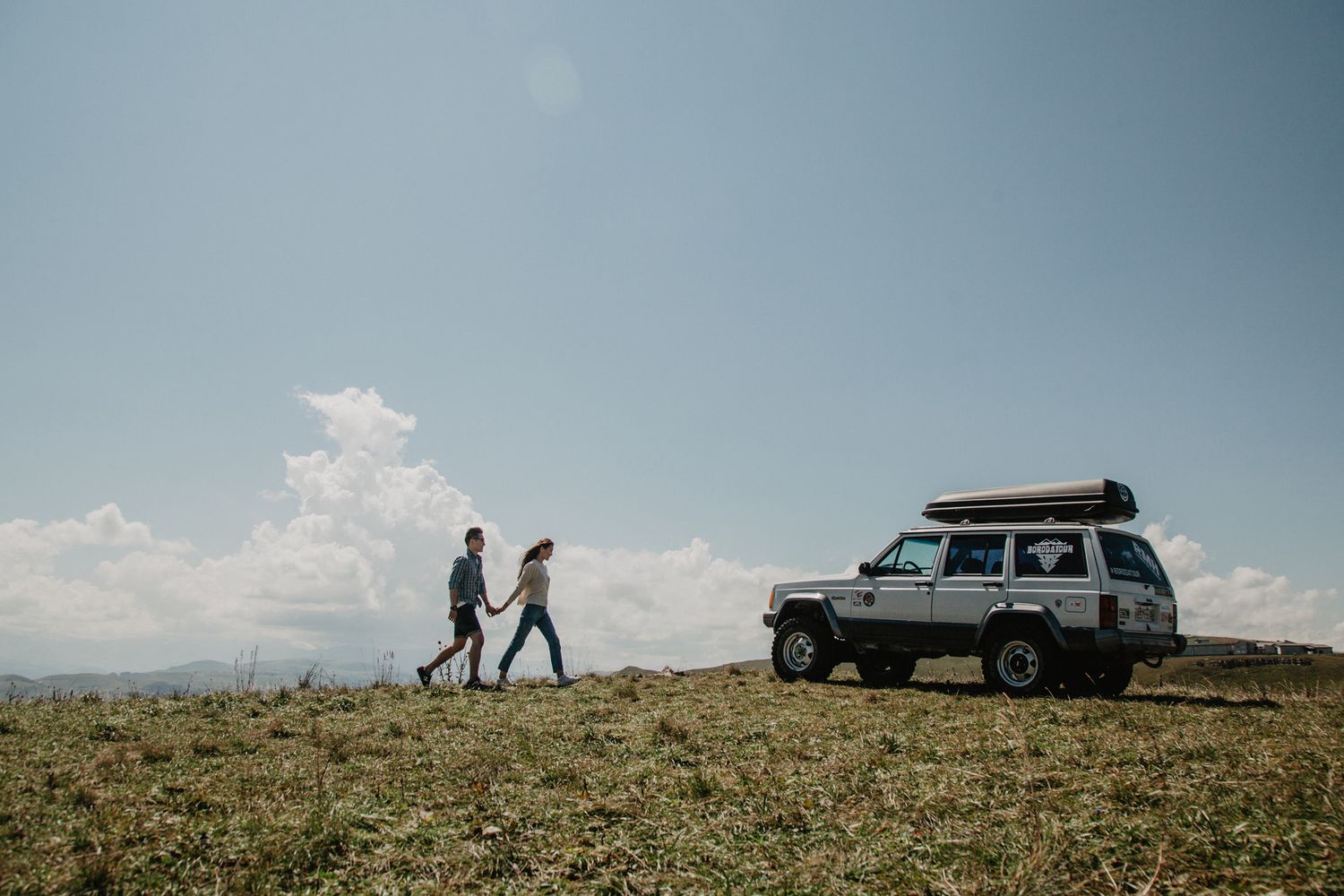 Two people walking near a parked SUV on a hilltop overlook.