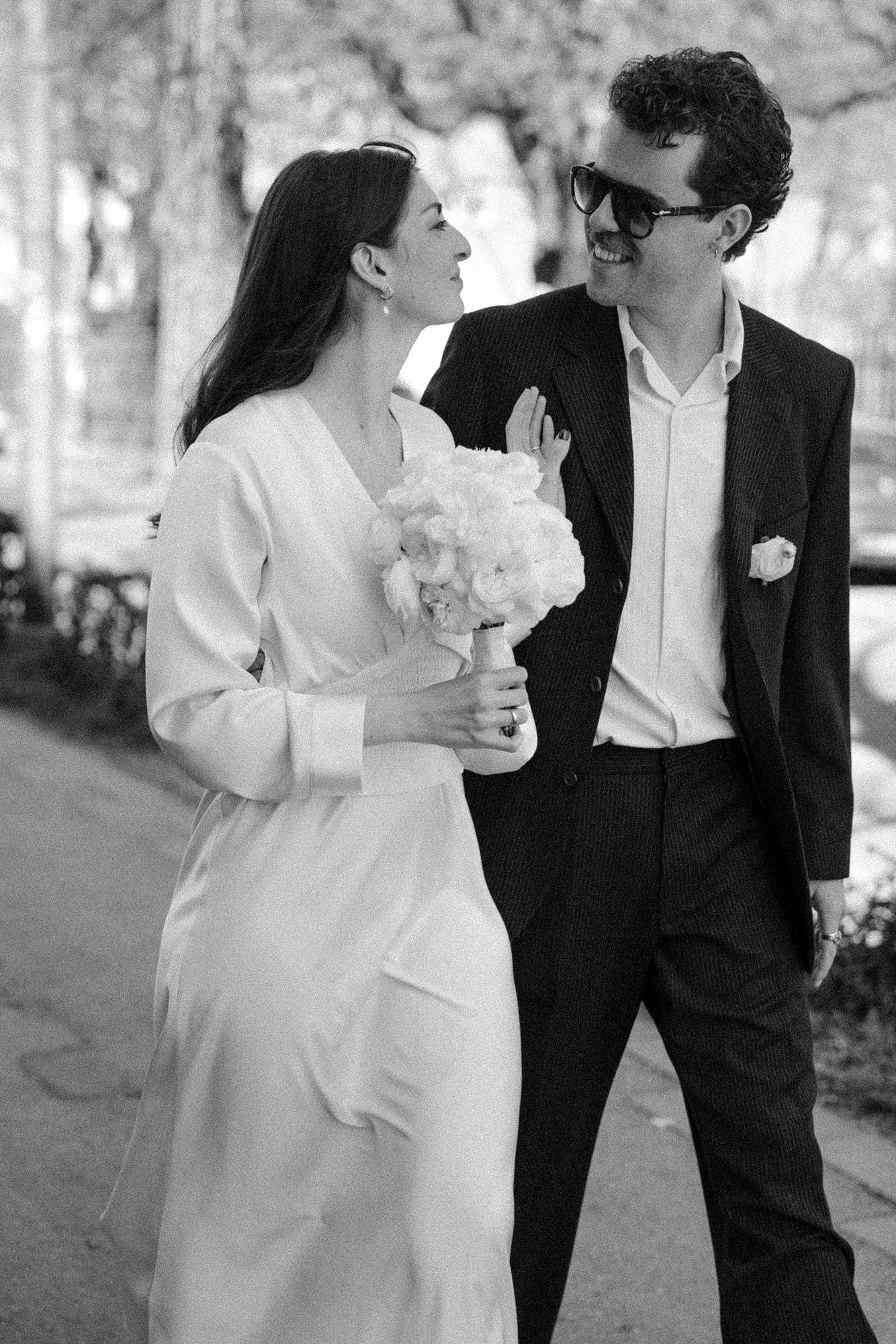 A black and white portrait of a couple sharing a joyful moment outdoors while holding a white bouquet.