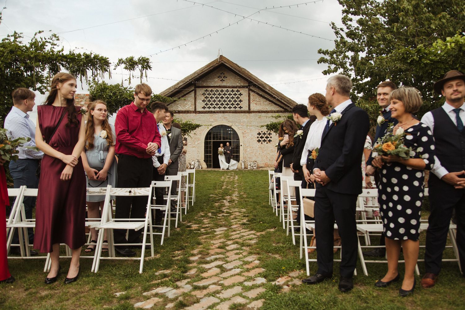 Outdoor wedding ceremony setup with white chairs on a lawn leading to a rustic brick building.