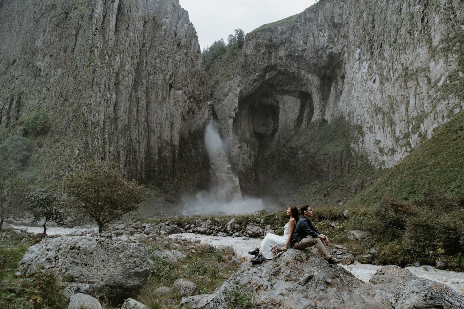 Wide angle view of couple sitting on rocks near majestic waterfall in limestone canyon.