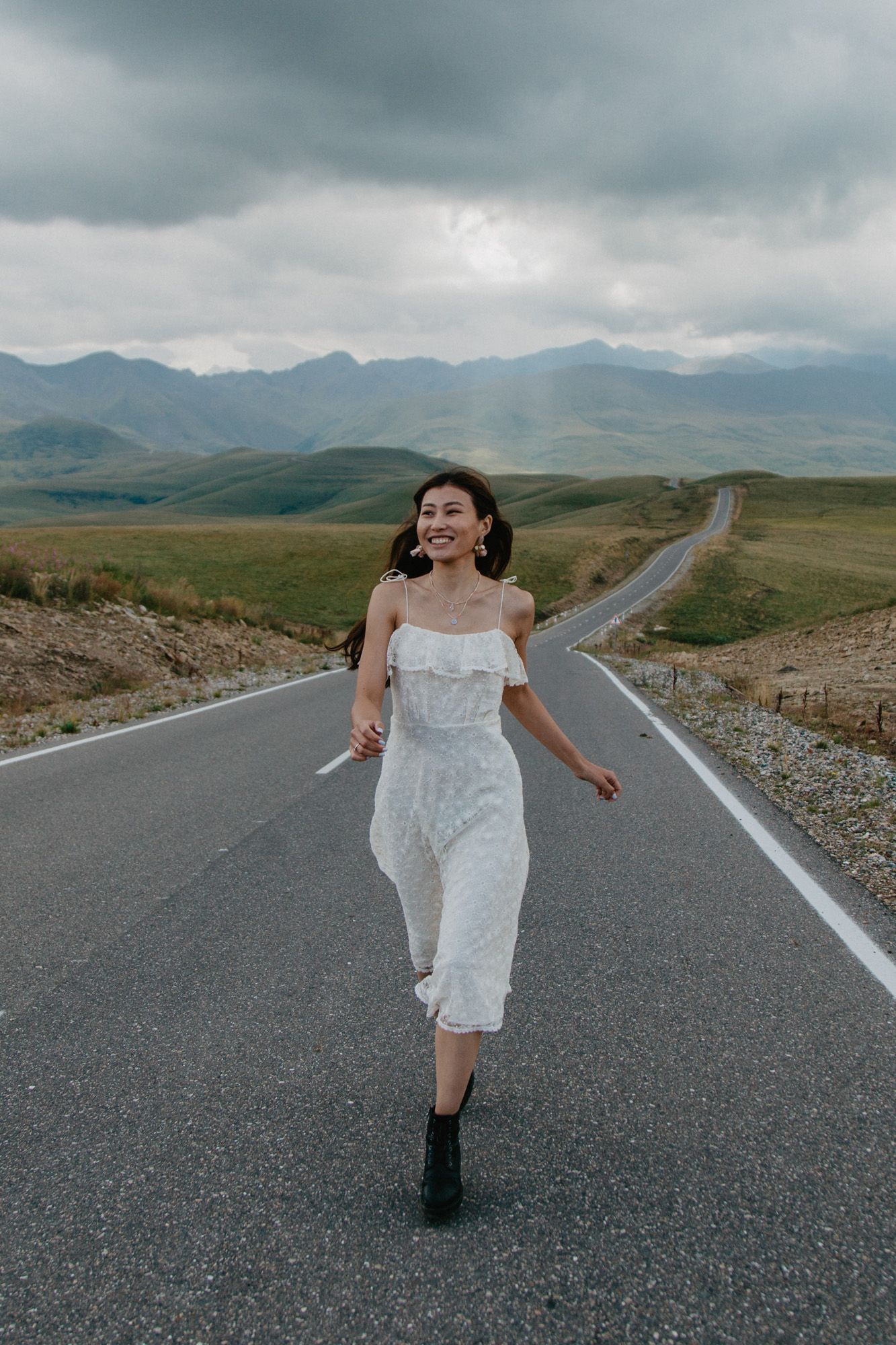 A figure in a white dress dances joyfully on a winding mountain road under stormy skies.