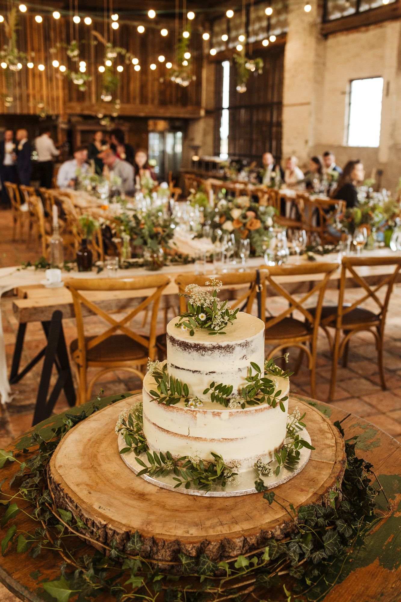 Three-tiered white wedding cake decorated with greenery on wooden slice in rustic venue.