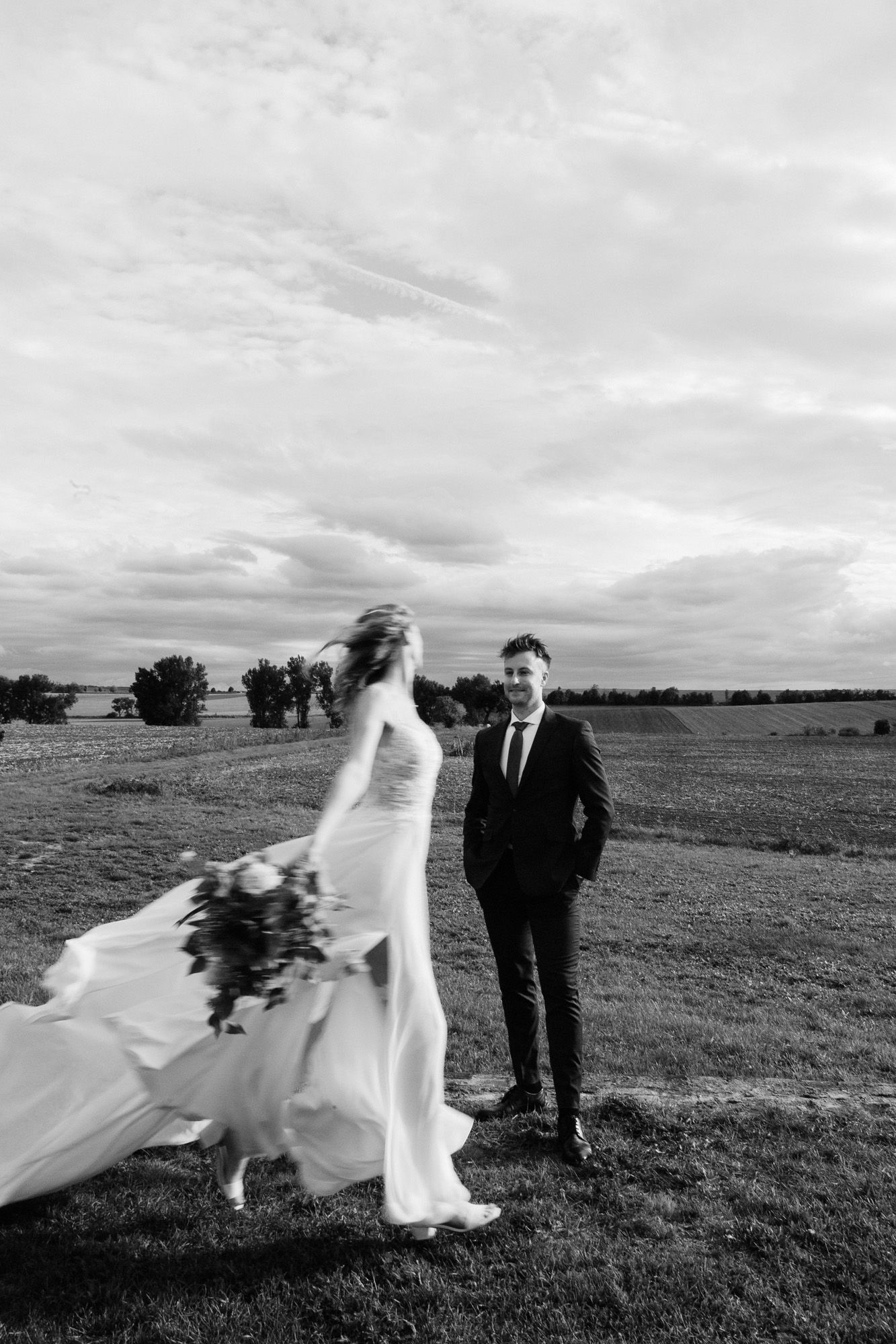 Dramatic black and white photo of newlyweds in an open field with the bride's dress catching wind.