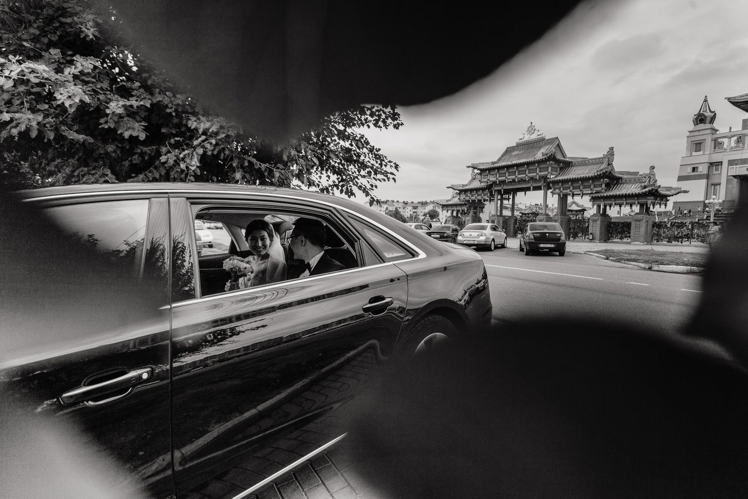 Black and white photo of car driving past traditional Asian buildings on street.