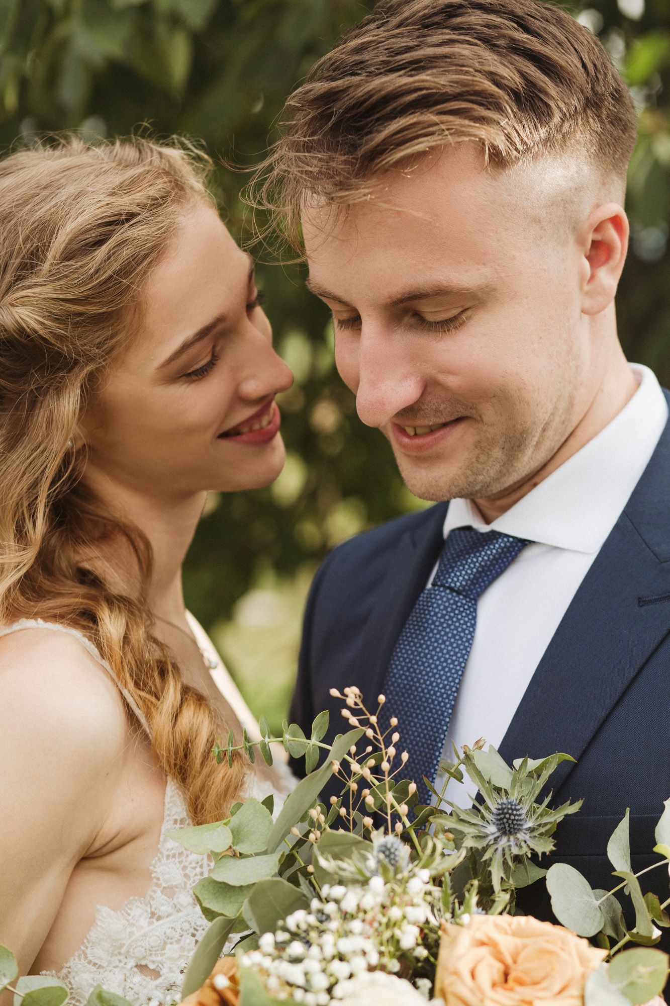 A romantic wedding couple shares an intimate moment outdoors with a bridal bouquet of peach roses and greenery.