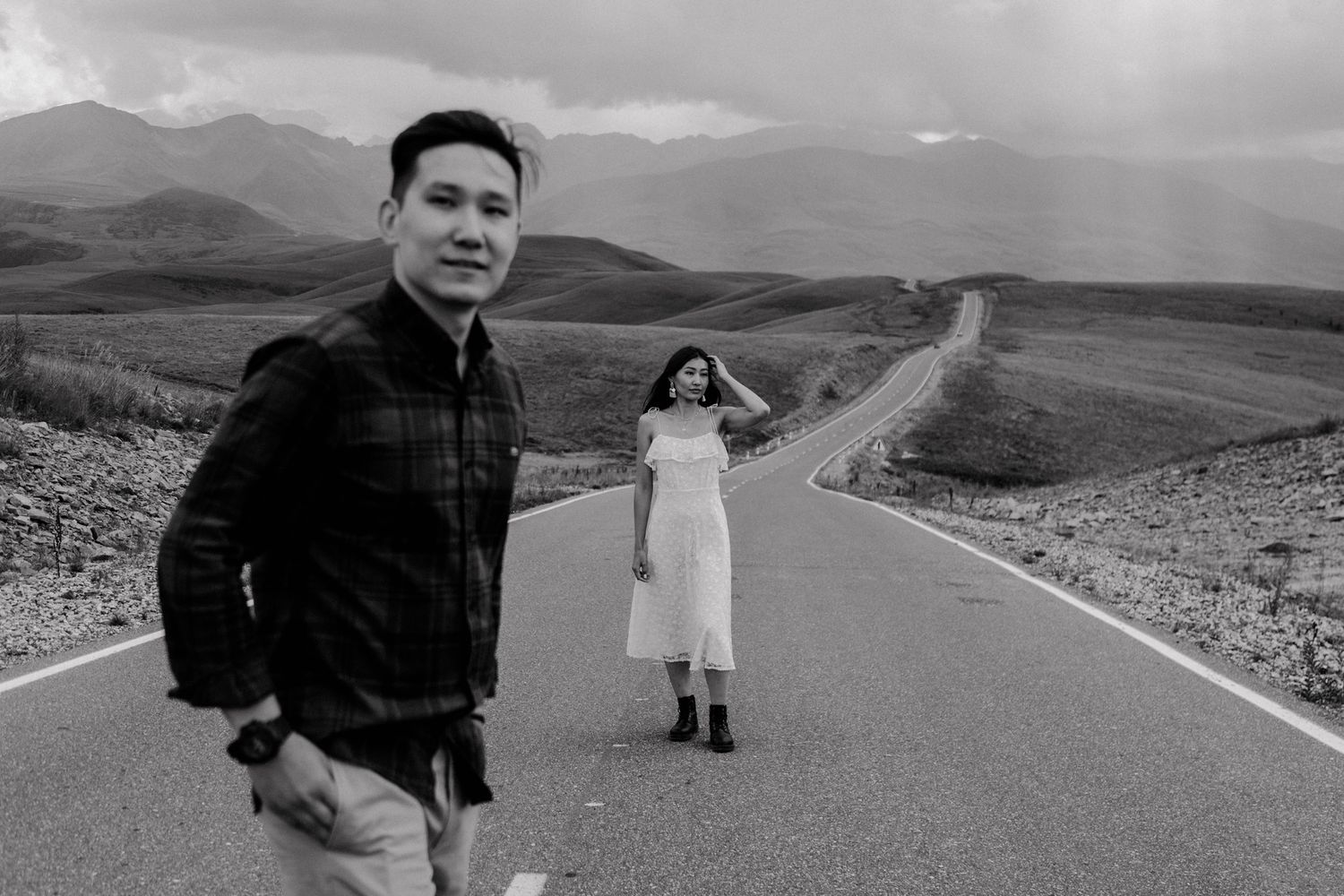 A couple poses creatively on a winding mountain road with scenic mountain views in black and white.