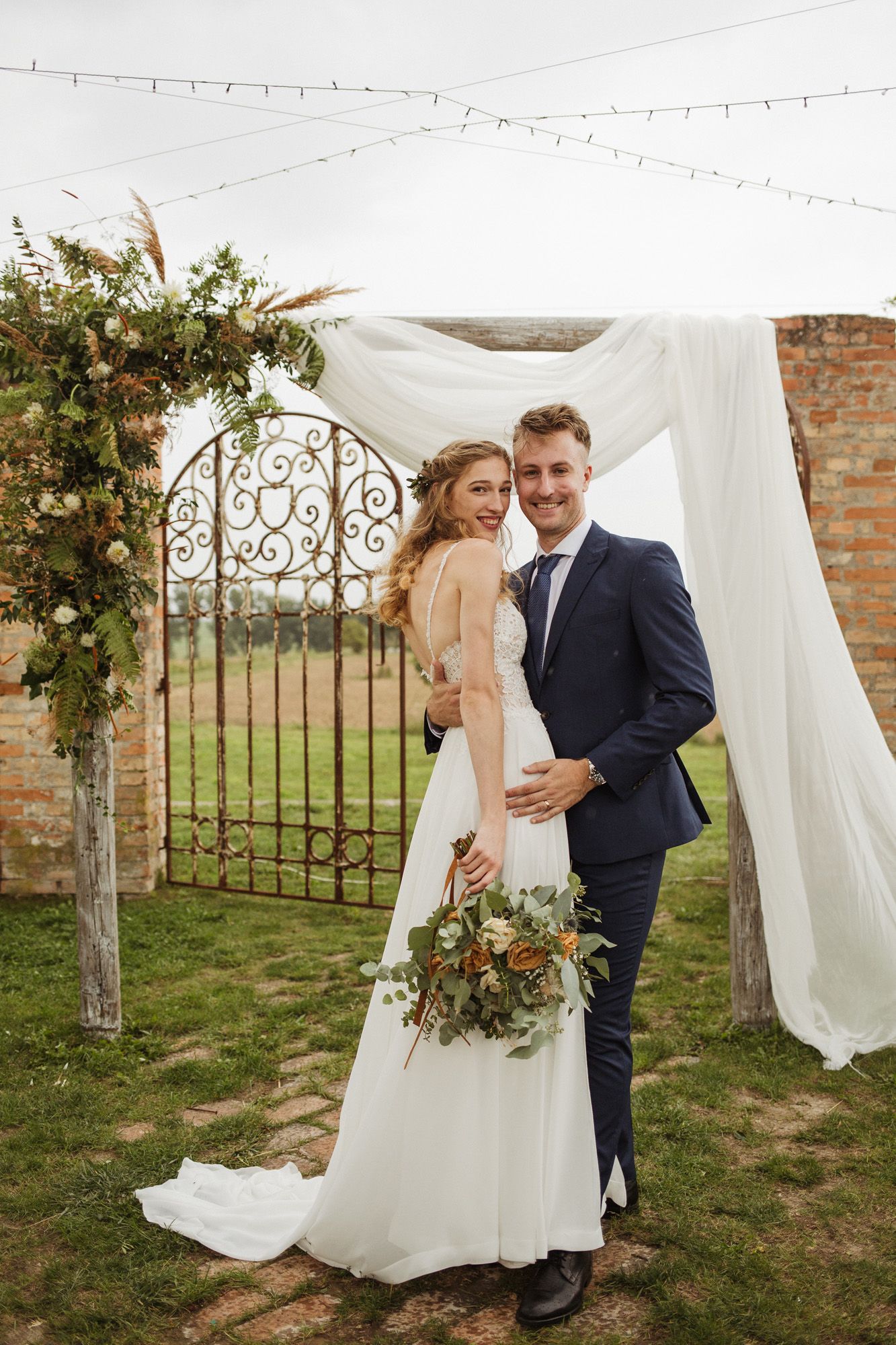 Outdoor wedding ceremony setup with white fabric draping and ornate metal arch decorated with greenery.
