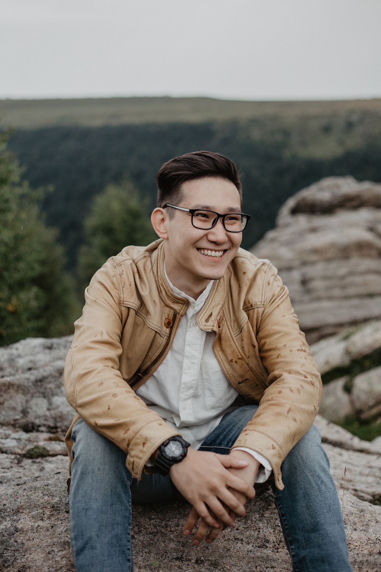 Person in tan jacket and glasses sitting on rocky terrain with mountains in background.