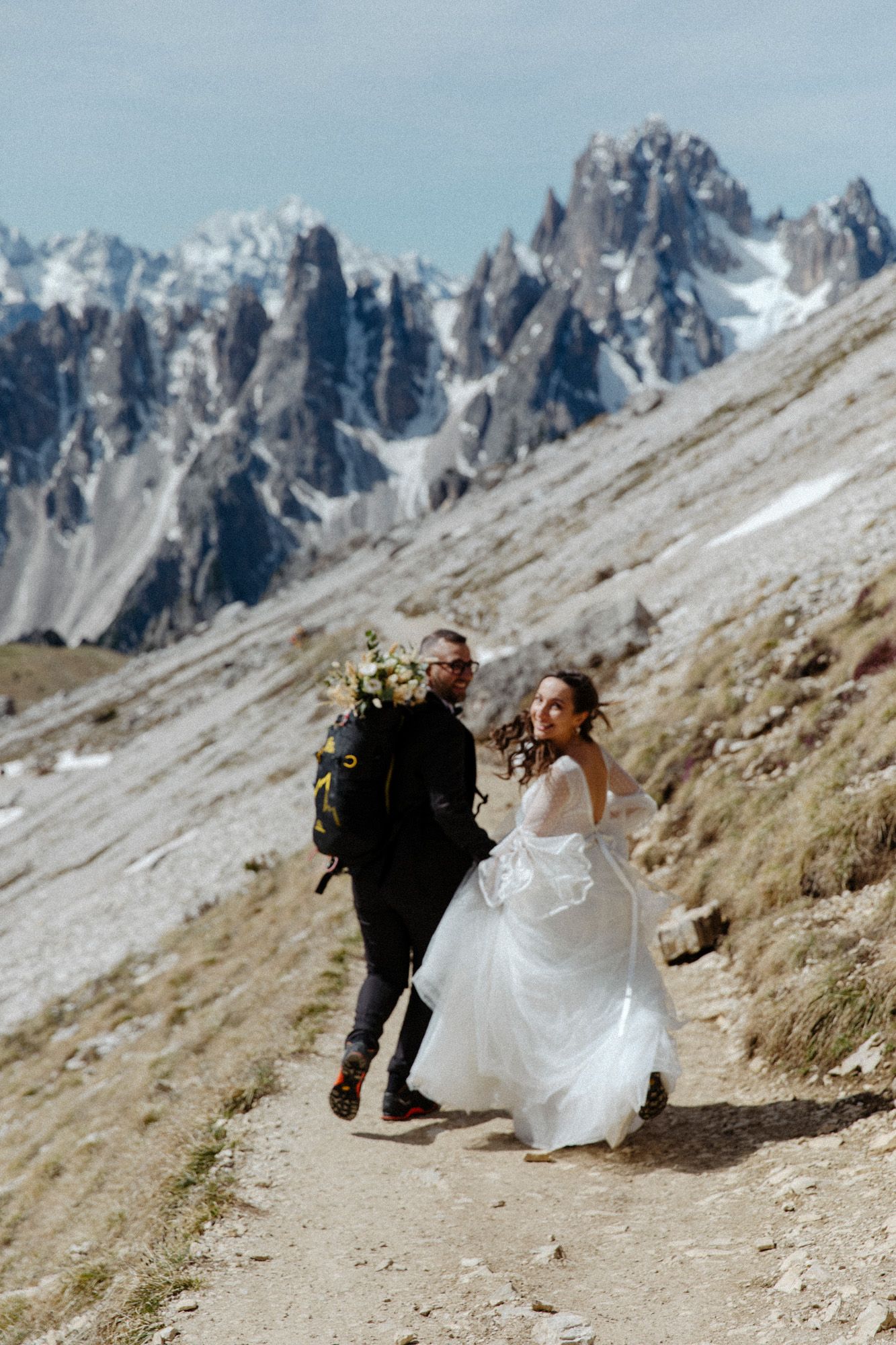 Adventure wedding couple hiking mountain trail with jagged peaks in background.