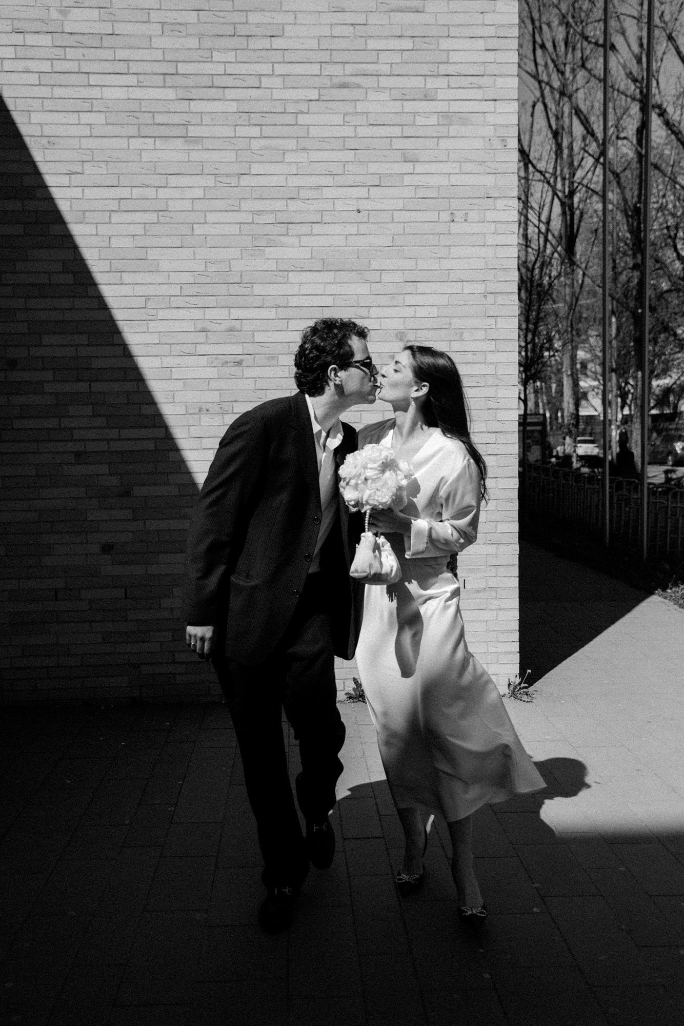 A black and white photo of a couple sharing an intimate moment against a brick wall with dramatic shadows.